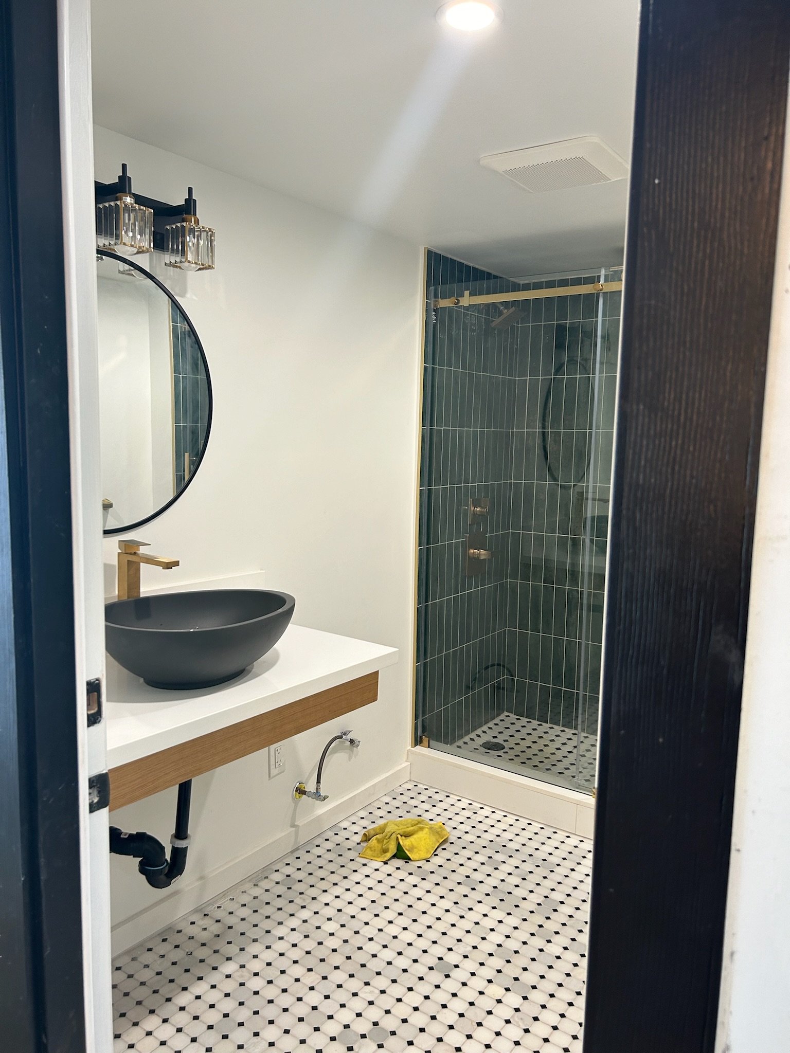 A bathroom with black and white patterned floor tiles, a small round black vessel sink on a white countertop with a wooden drawer, and a shower with dark green tile walls and a brass sliding door rail.