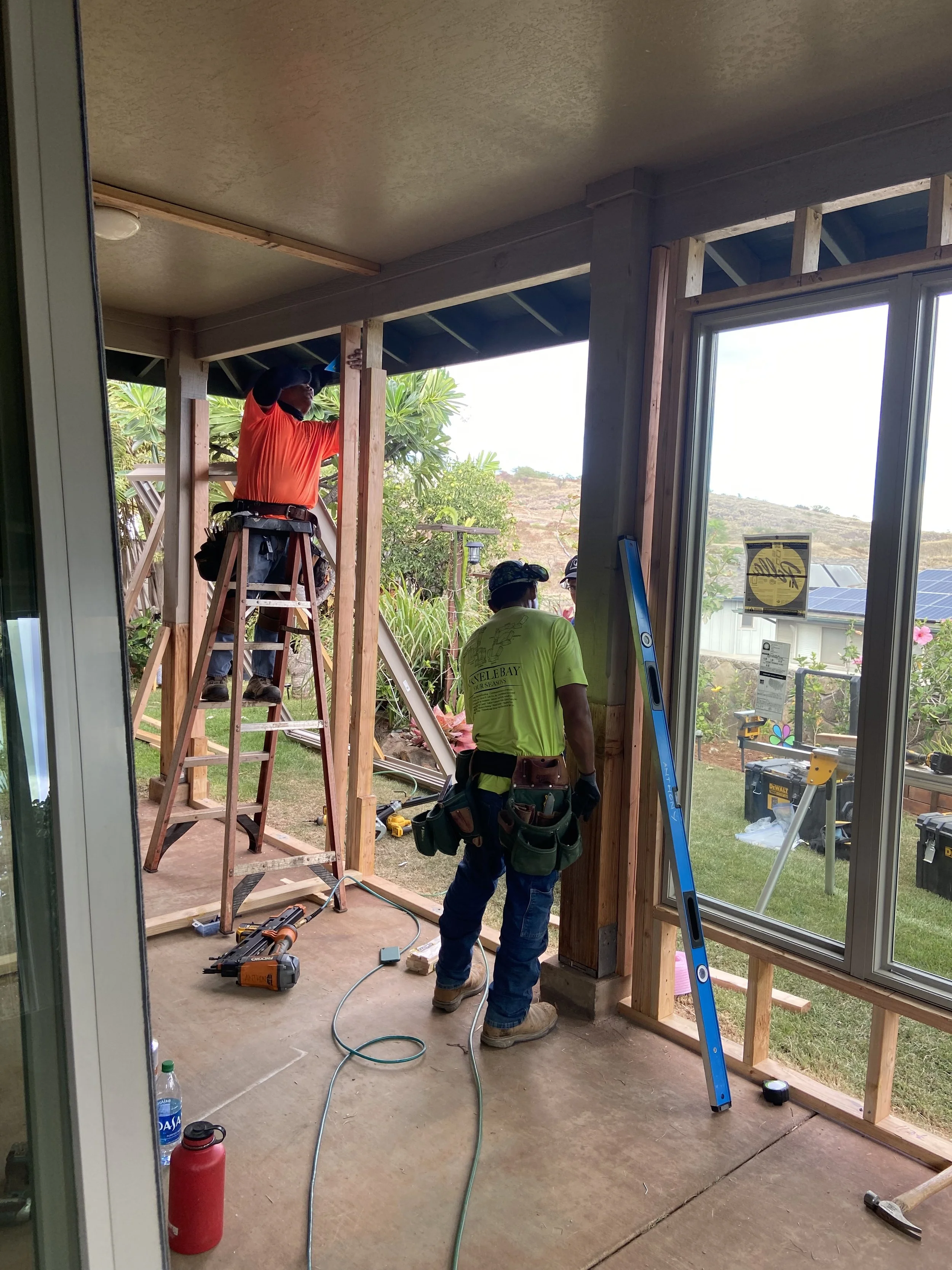 Home construction site with two workers installing a wooden frame for a porch or deck. One worker is on a ladder working on the roof, while the other stands on the ground with a tool belt. Construction tools and equipment are scattered on the floor, and there is a view of the outdoors with greenery and a house with solar panels in the background.