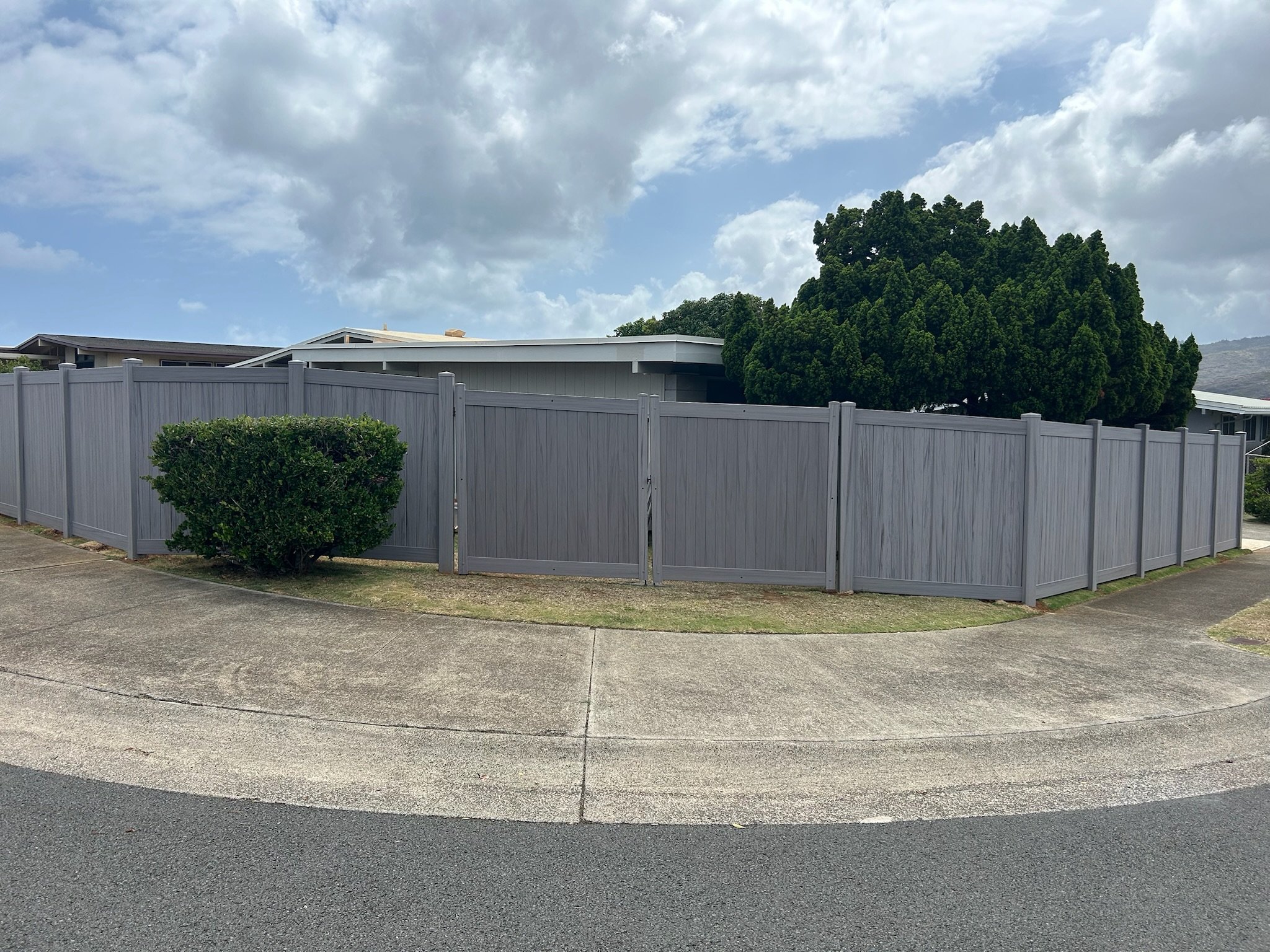 A view of a residential neighborhood with a gray fence, a bush, a large tree, and part of a house with a sloped roof under a partly cloudy sky.