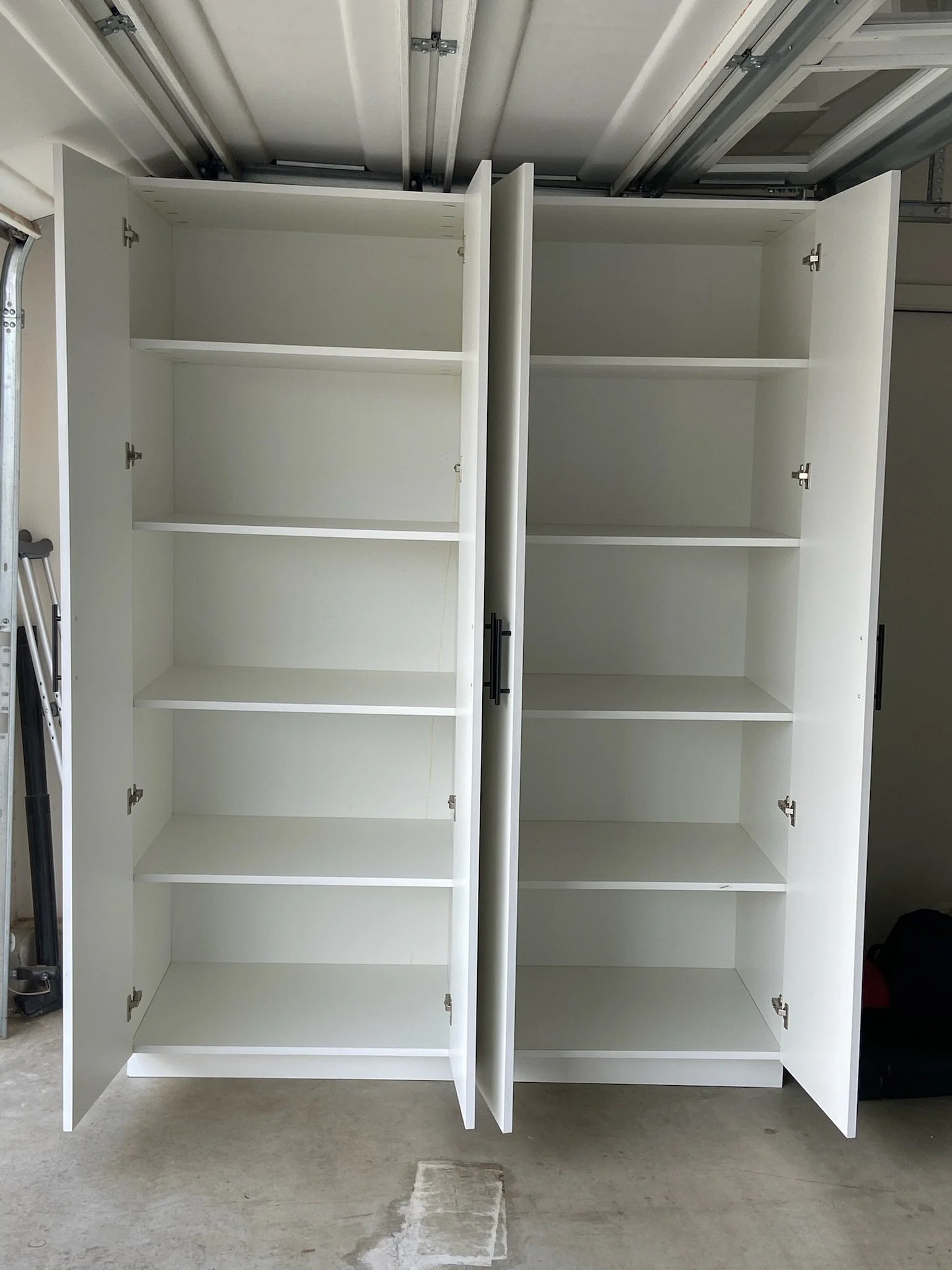 Empty white kitchen cabinet with glass panel doors and open shelves in a garage setting.