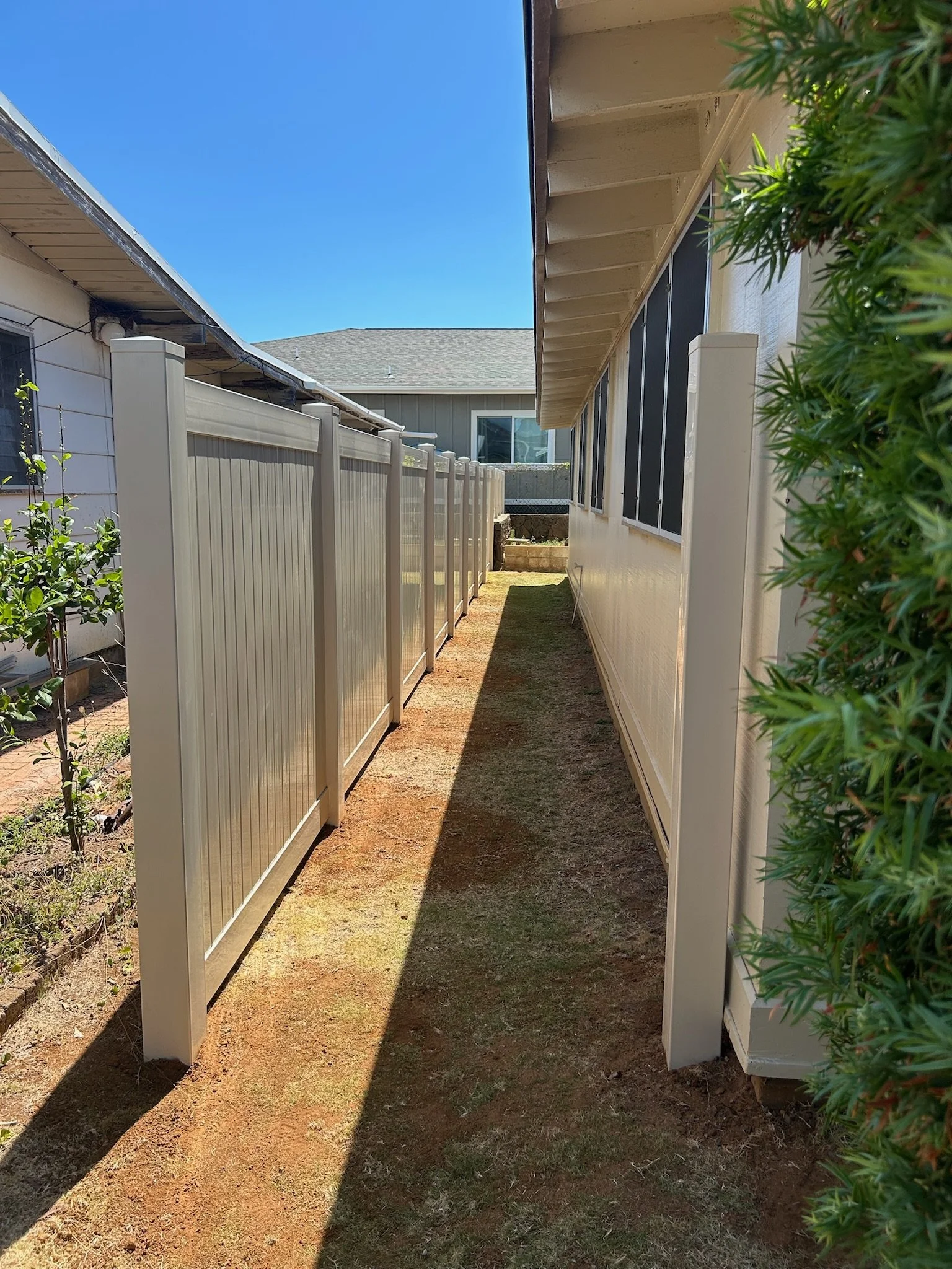 A narrow dirt pathway between two houses, with a beige fence on the left and a beige house with large black window screens on the right, and a clear blue sky overhead.