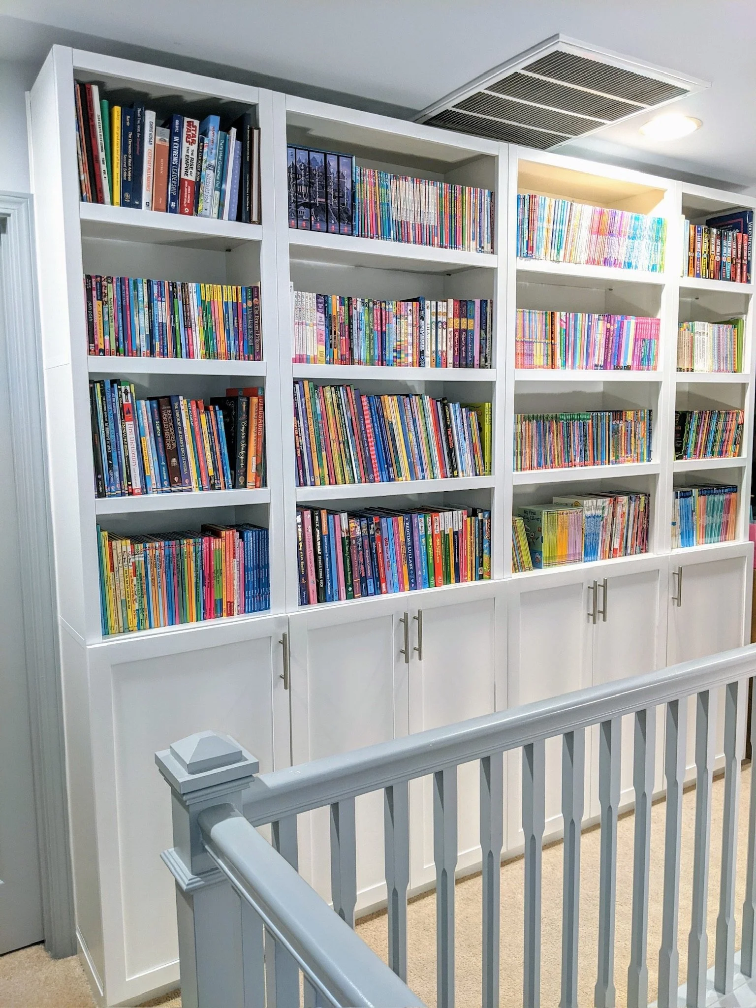 A white bookshelf filled with colorful books, located in an indoor space with beige carpet, white baseboards, and a gray ceiling with a vent and a ceiling light.