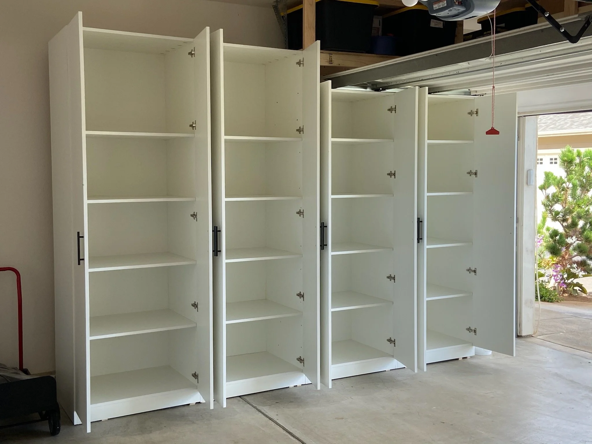 White empty shelving units arranged in a garage, with some doors open showing interior shelves, partially enclosed by a wall and next to a window with greenery outside.