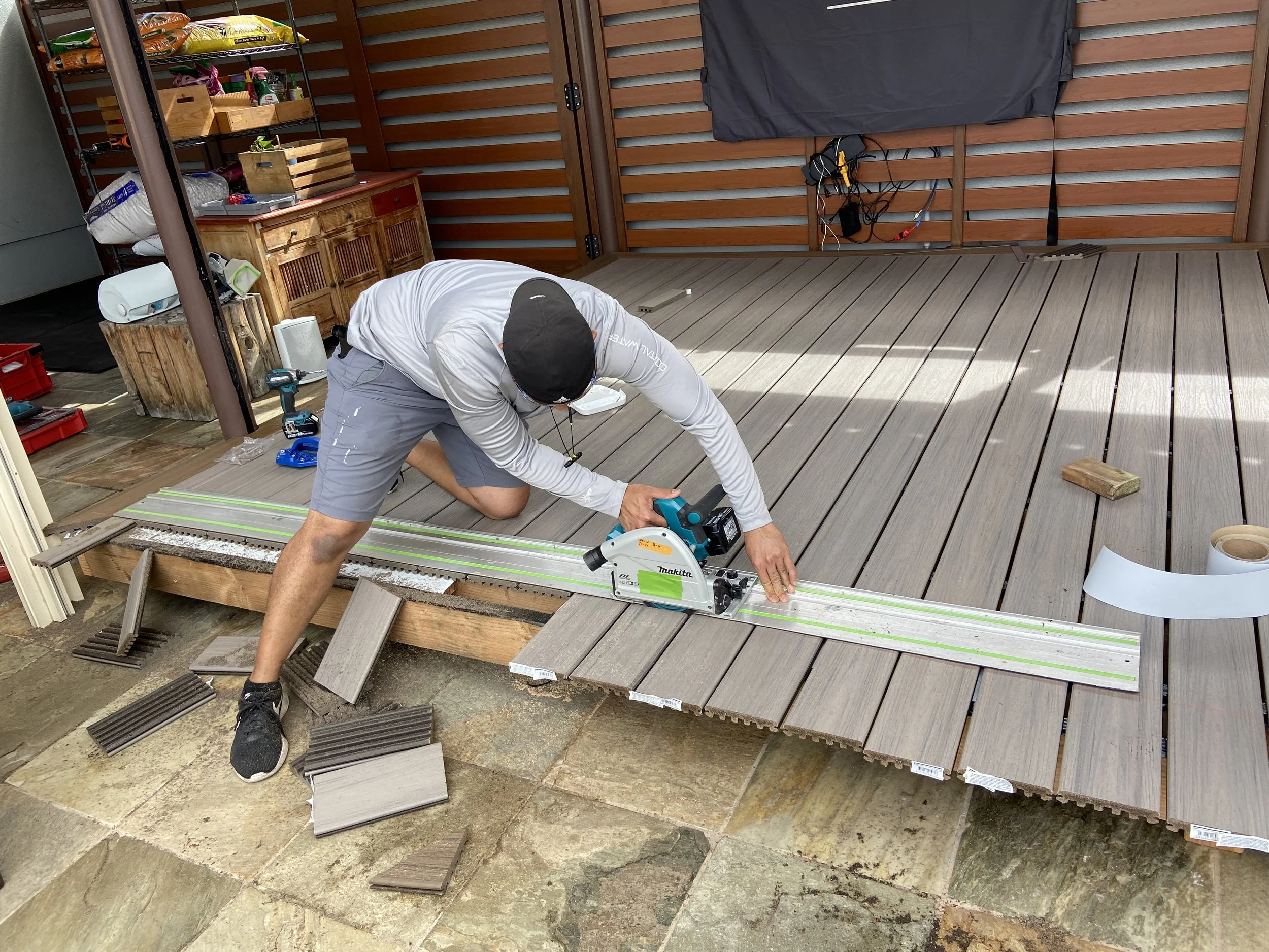 Person installing or repairing outdoor deck flooring with power tools, surrounded by construction materials and tools.