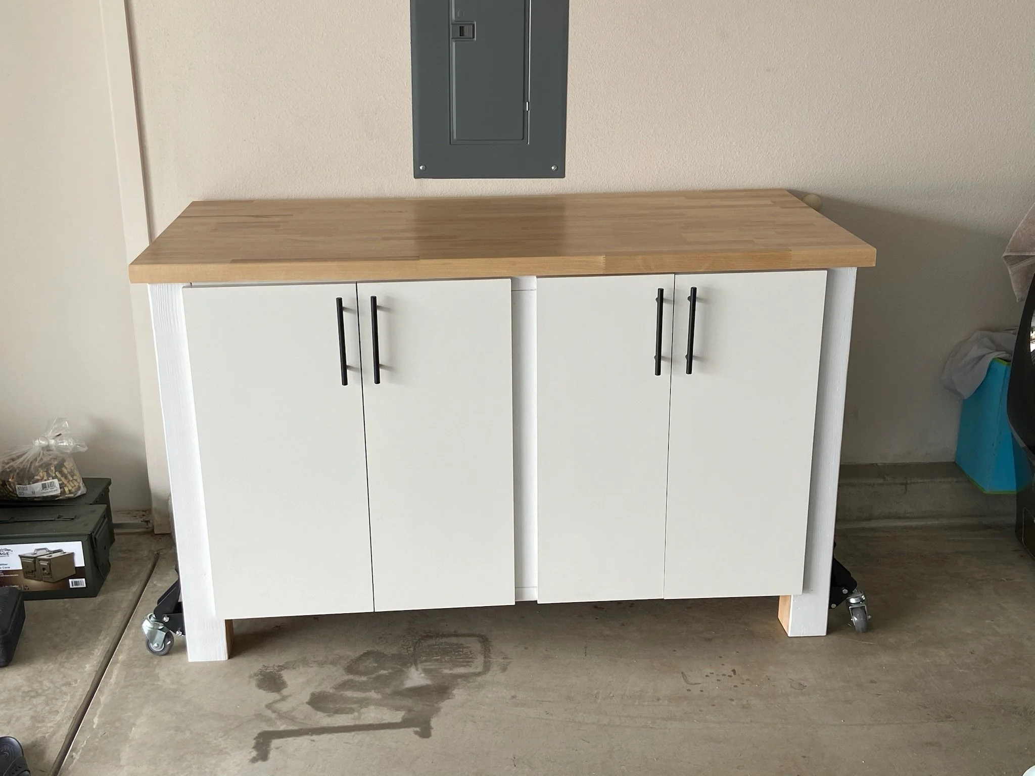 A white cabinet with a natural wood top on wheels, positioned against a beige wall. There is an electrical panel on the wall above the cabinet, and various items including a plastic bag with food, a small black container, and a blue recycling bin are