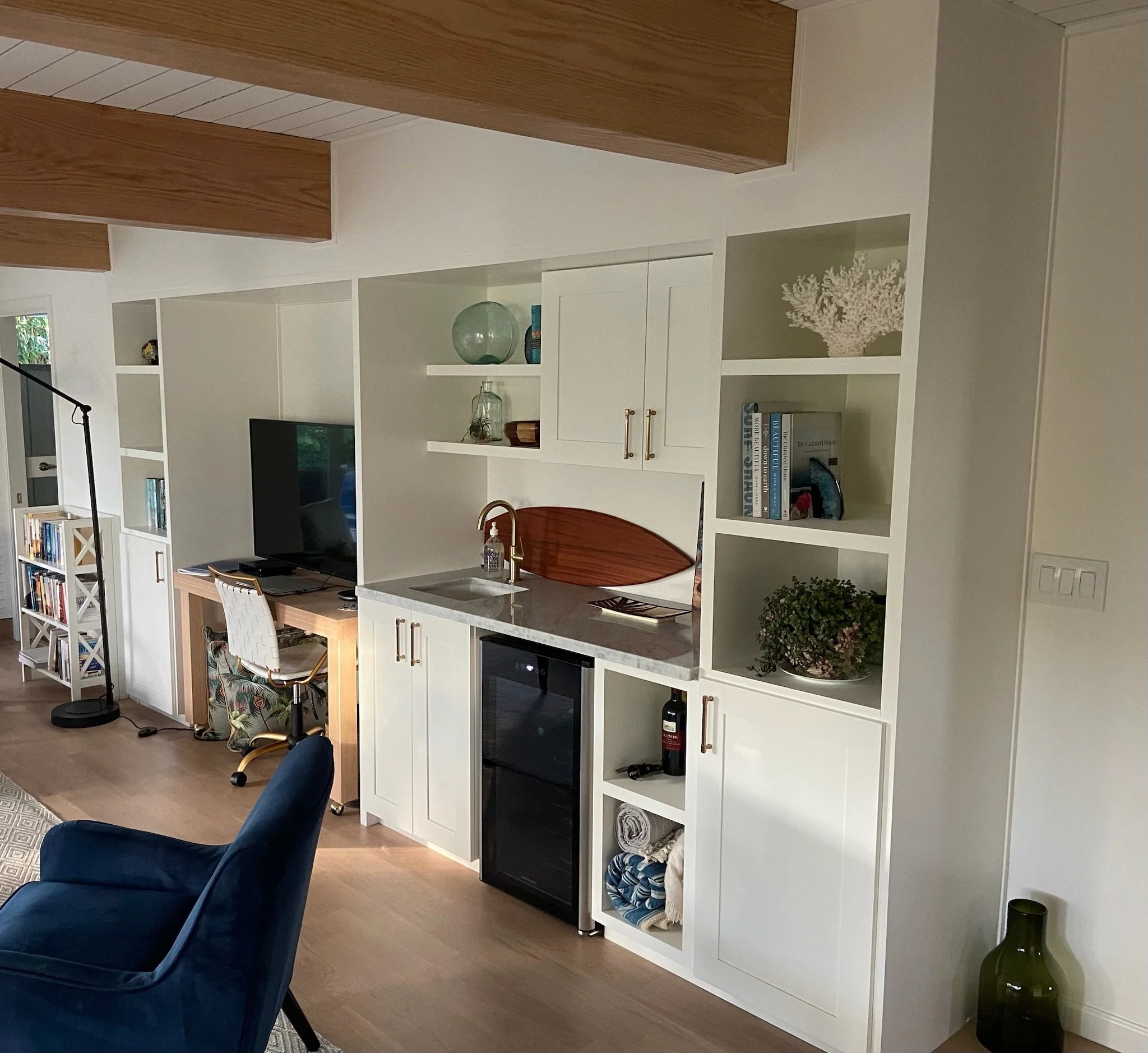 Living room area with a built-in white bookshelf, a flat-screen TV, and decorative items like plants and books. There are navy chairs, a wooden table, and a wood ceiling with visible beams.