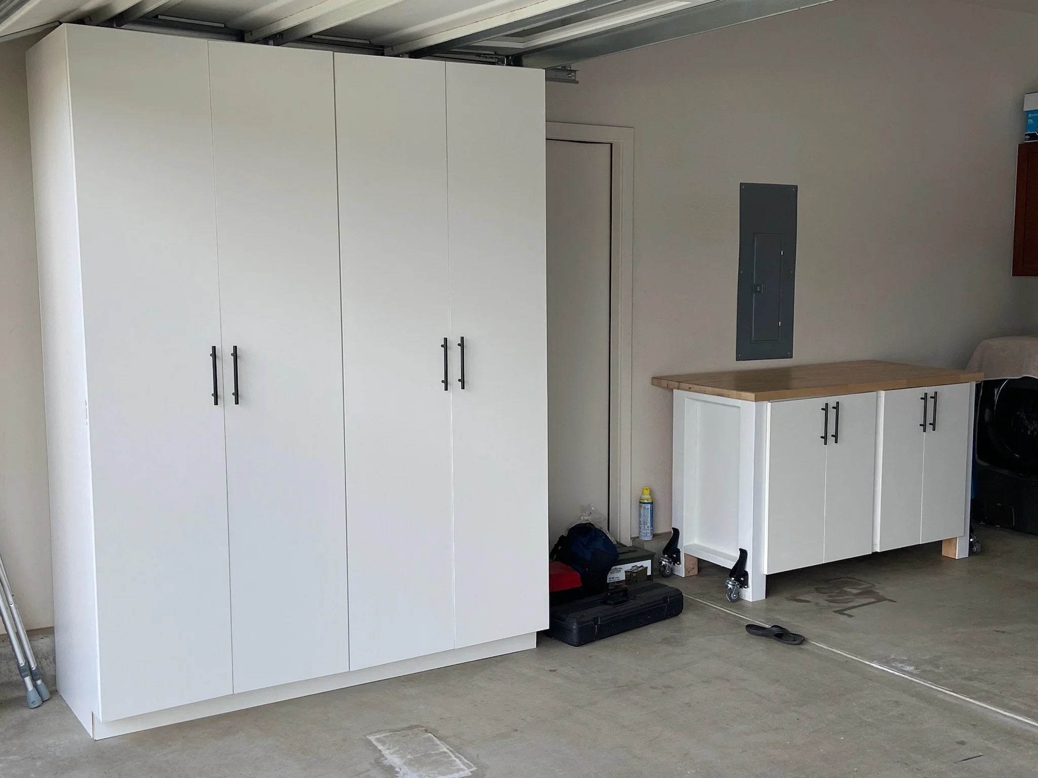 A white storage cabinet with black handles in a garage. To the right, there is a white cabinet on wheels, a wooden countertop, and various tools and items underneath and beside it. The garage wall has electrical panels and a small cabinet.