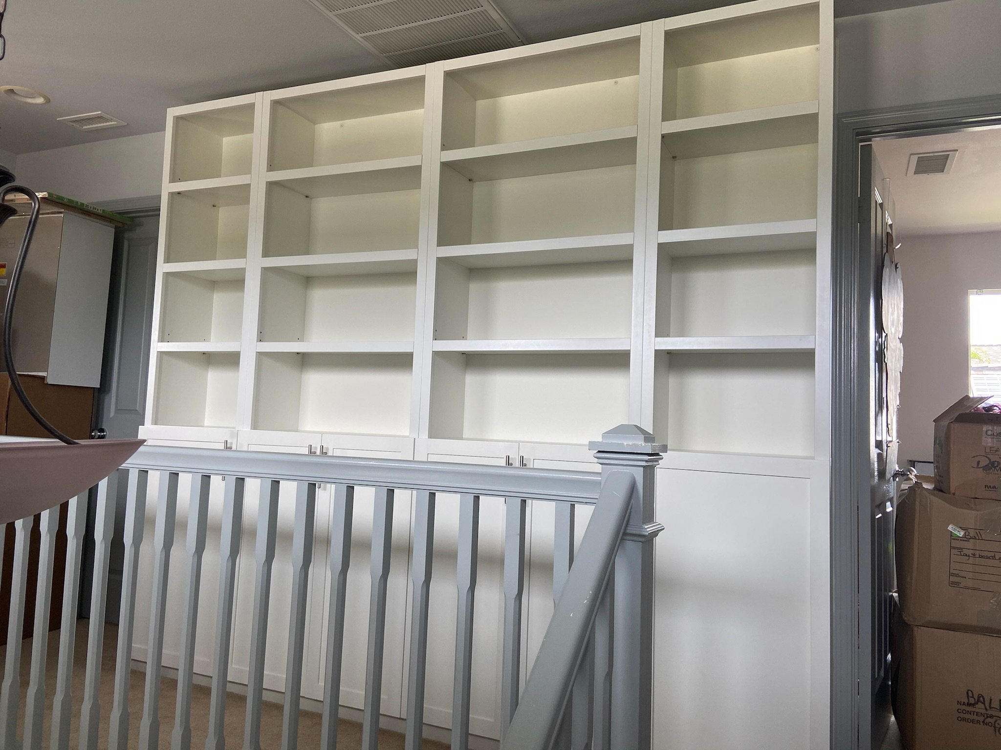 Empty white bookshelf with multiple shelves in a room, next to a staircase and cardboard boxes.