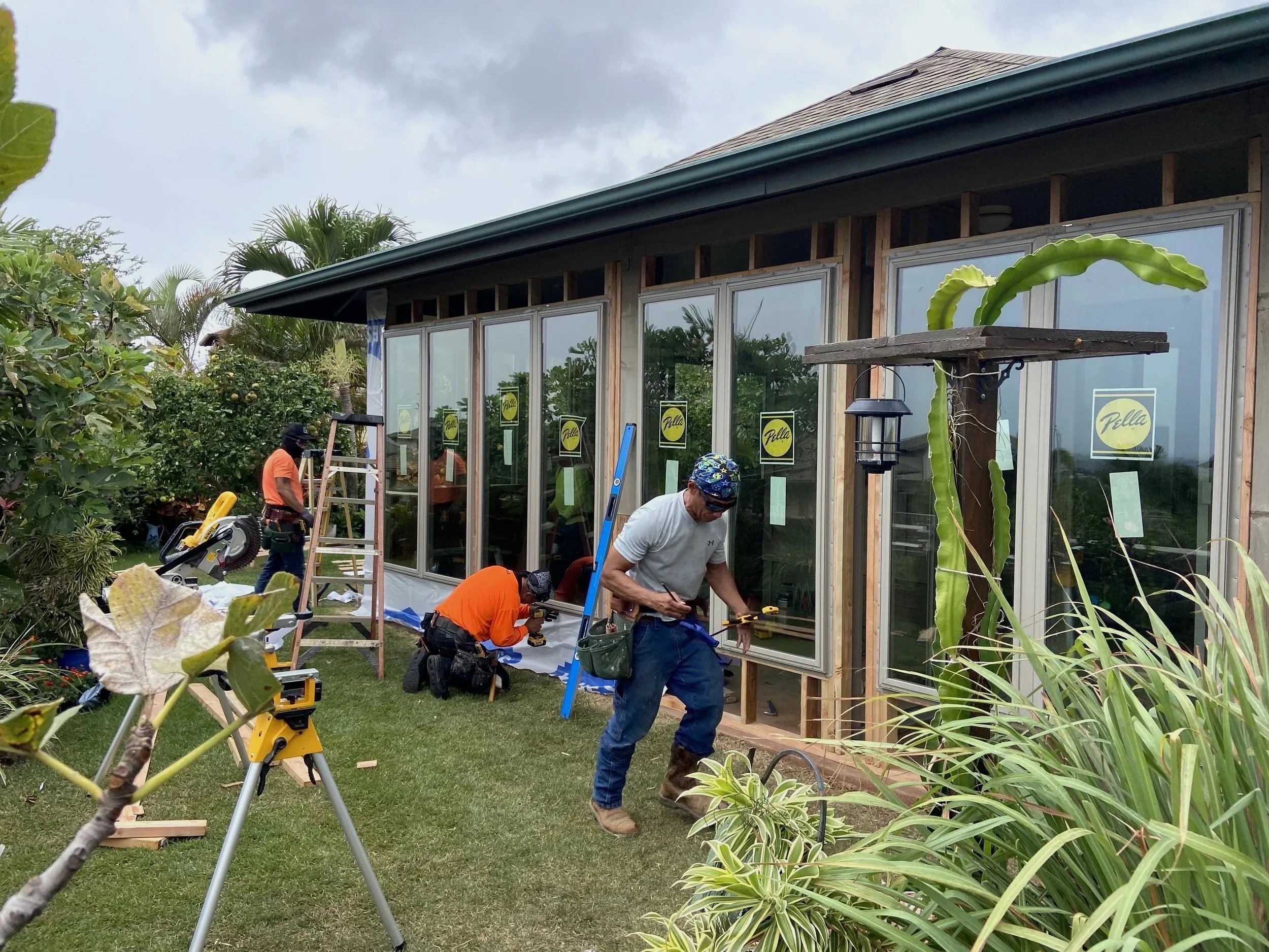 Construction workers installing large glass windows on a building under construction, surrounded by greenery on a cloudy day.