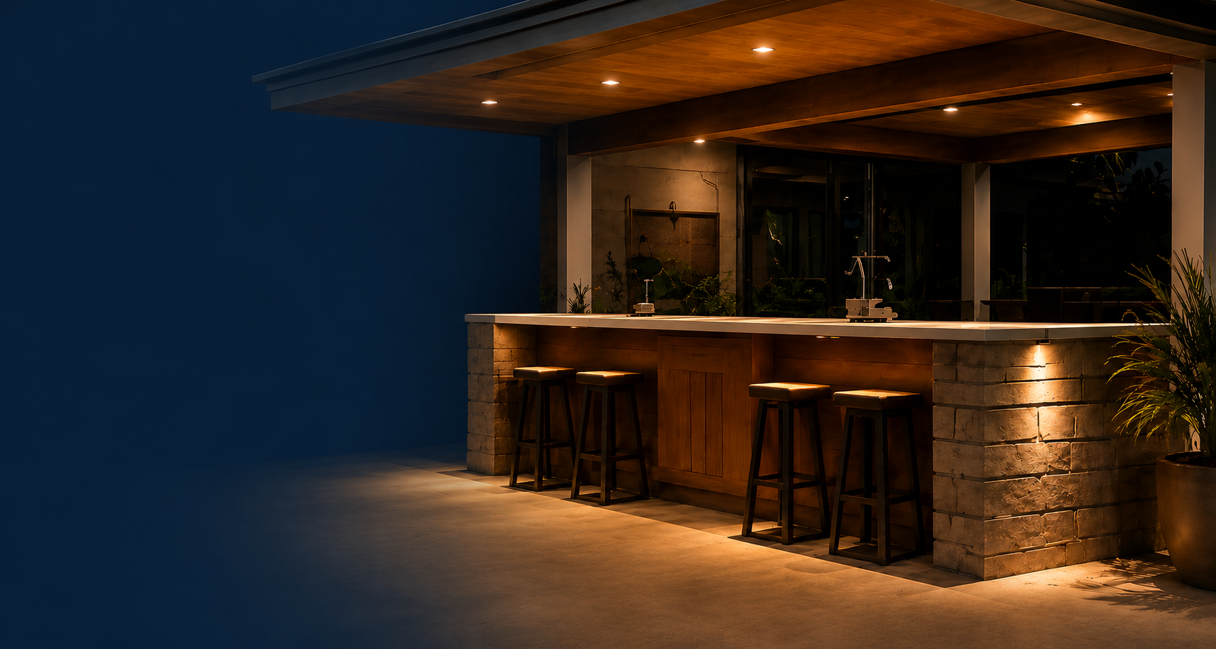 Modern outdoor bar area at night with four bar stools, a stone and wood counter, warm lighting, and plants in the background.