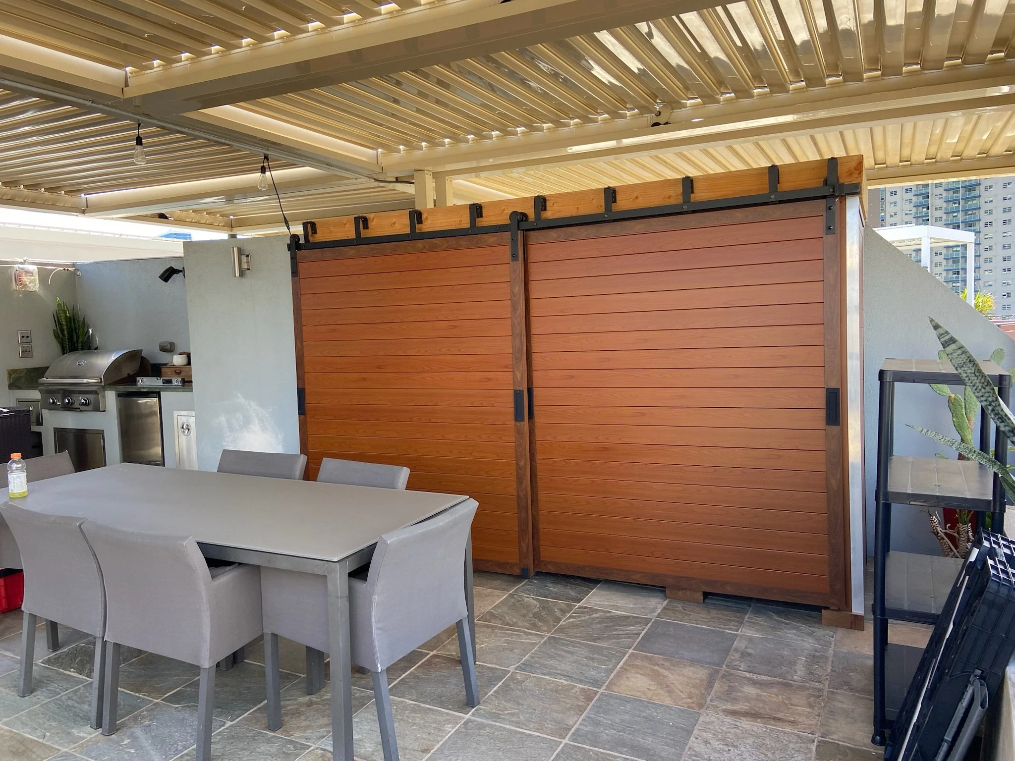 An outdoor patio area with a table and chairs, a wooden sliding door, a grill, and a partial view of high-rise buildings in the background.