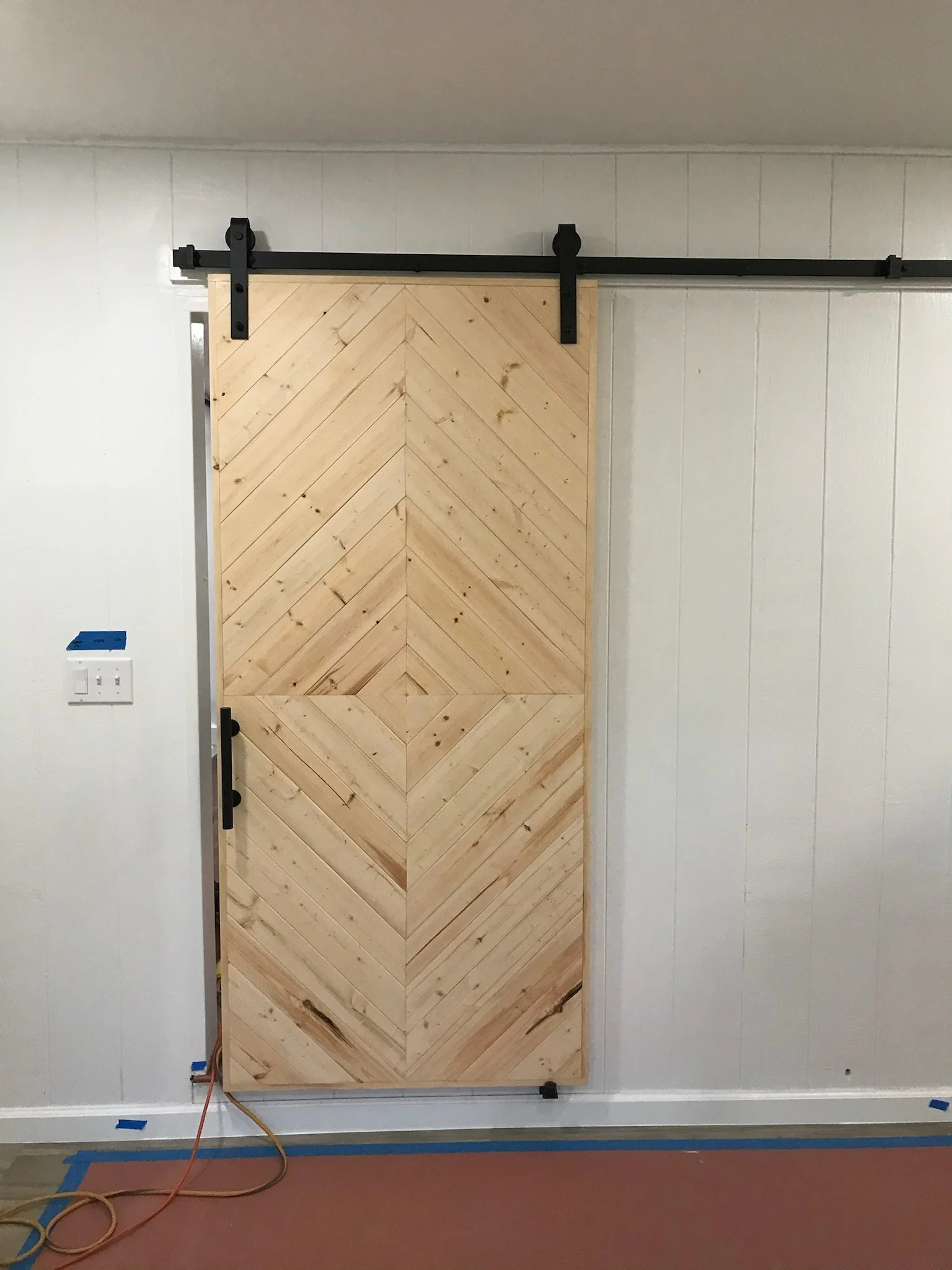 A wooden sliding barn door installed on a black metal track, with plastic wall protectors at the top and bottom, in a room with white paneled walls, electrical outlets, and construction equipment on the floor.