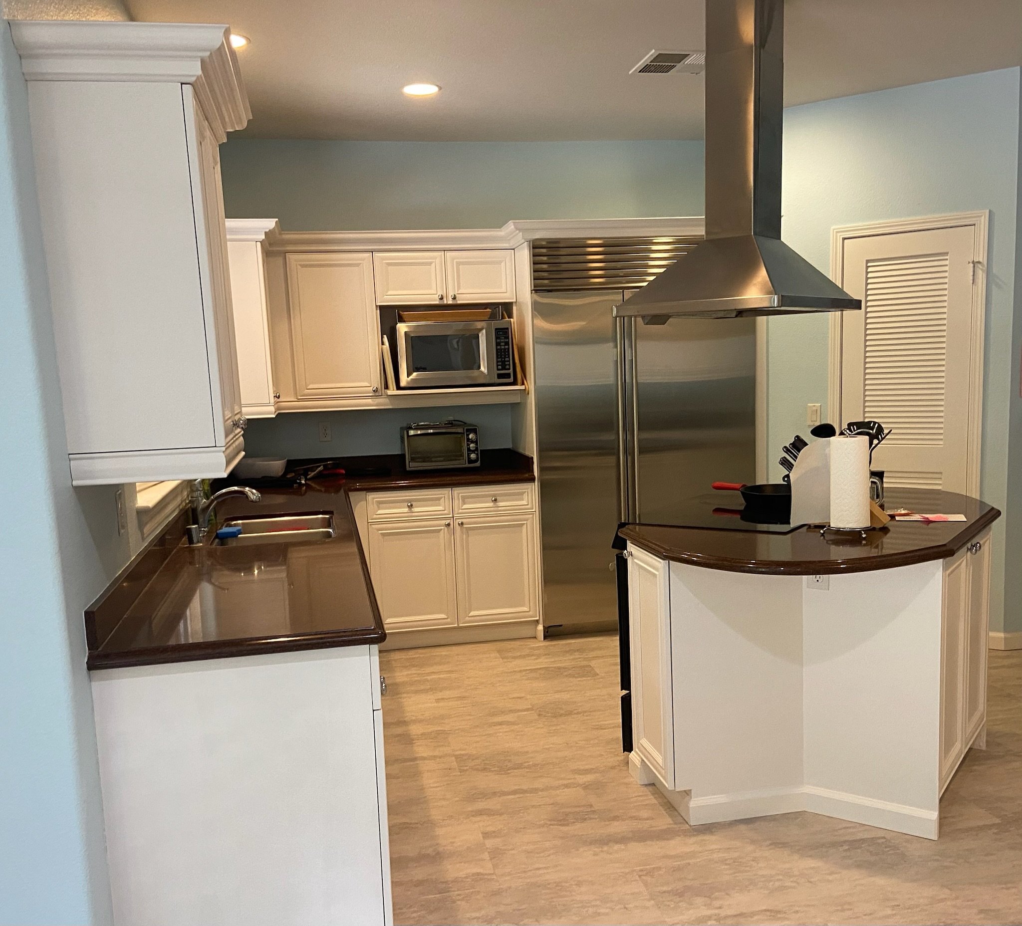 Kitchen with white cabinets, stainless steel range hood, built-in microwave, and small oven on the countertop. There are paper towels and kitchen utensils on the counter. Light teal walls and wood flooring. Part of a kitchen island is visible.