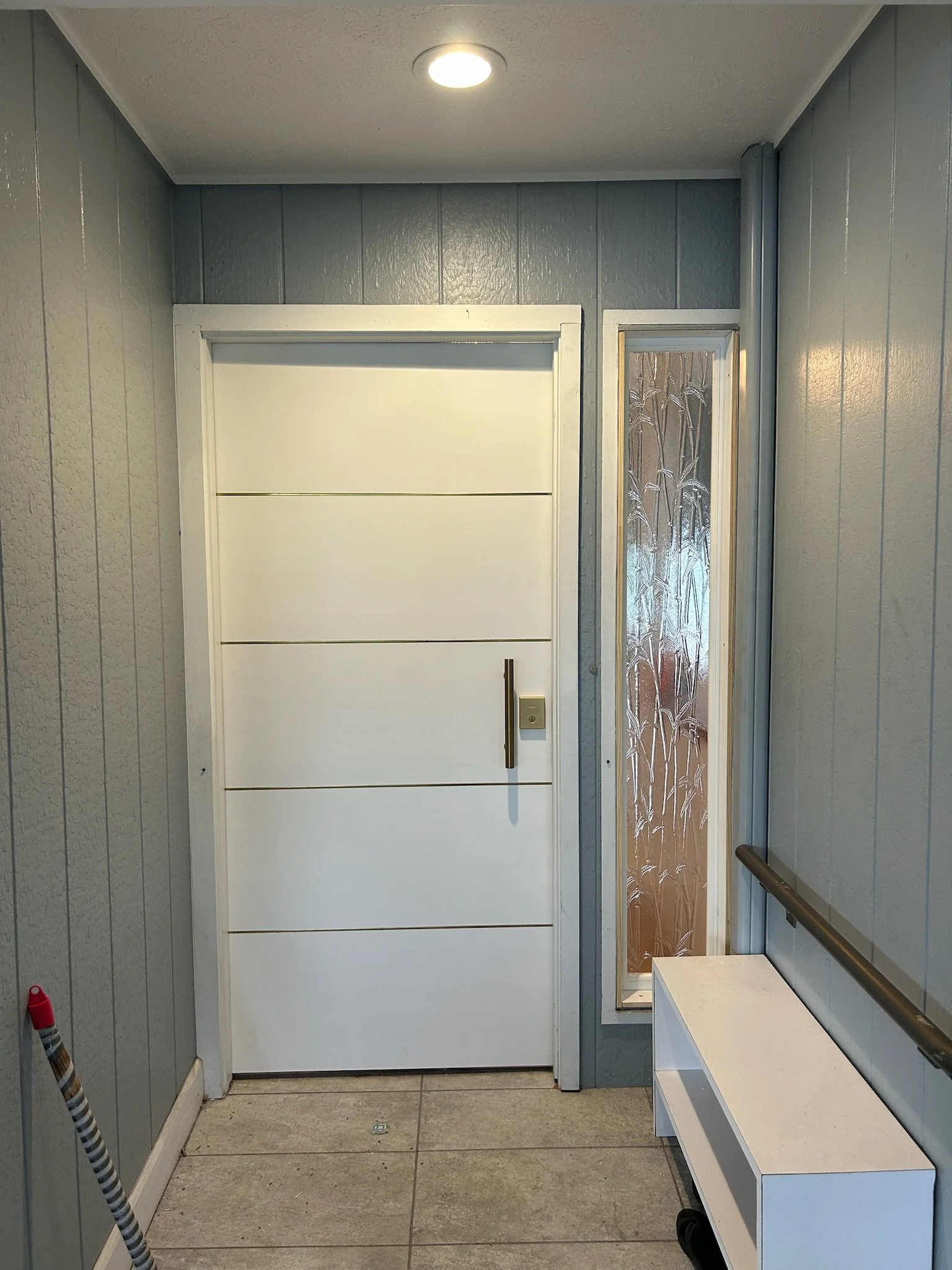 Indoor elevator lobby with gray wood panel walls, tiled floor, a white door, and a decorative glass window to the right of the door. There is a bench on the right side and a handrail on the right wall.
