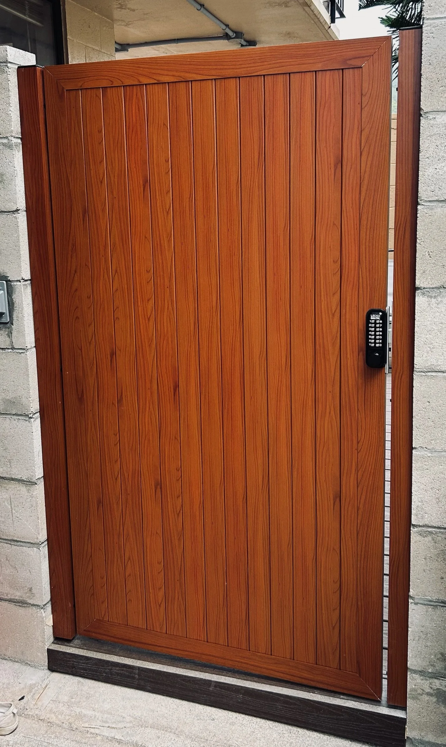 A wooden gate with vertical panels, a black lock on the right side, and brick columns on either side, situated outside a building.