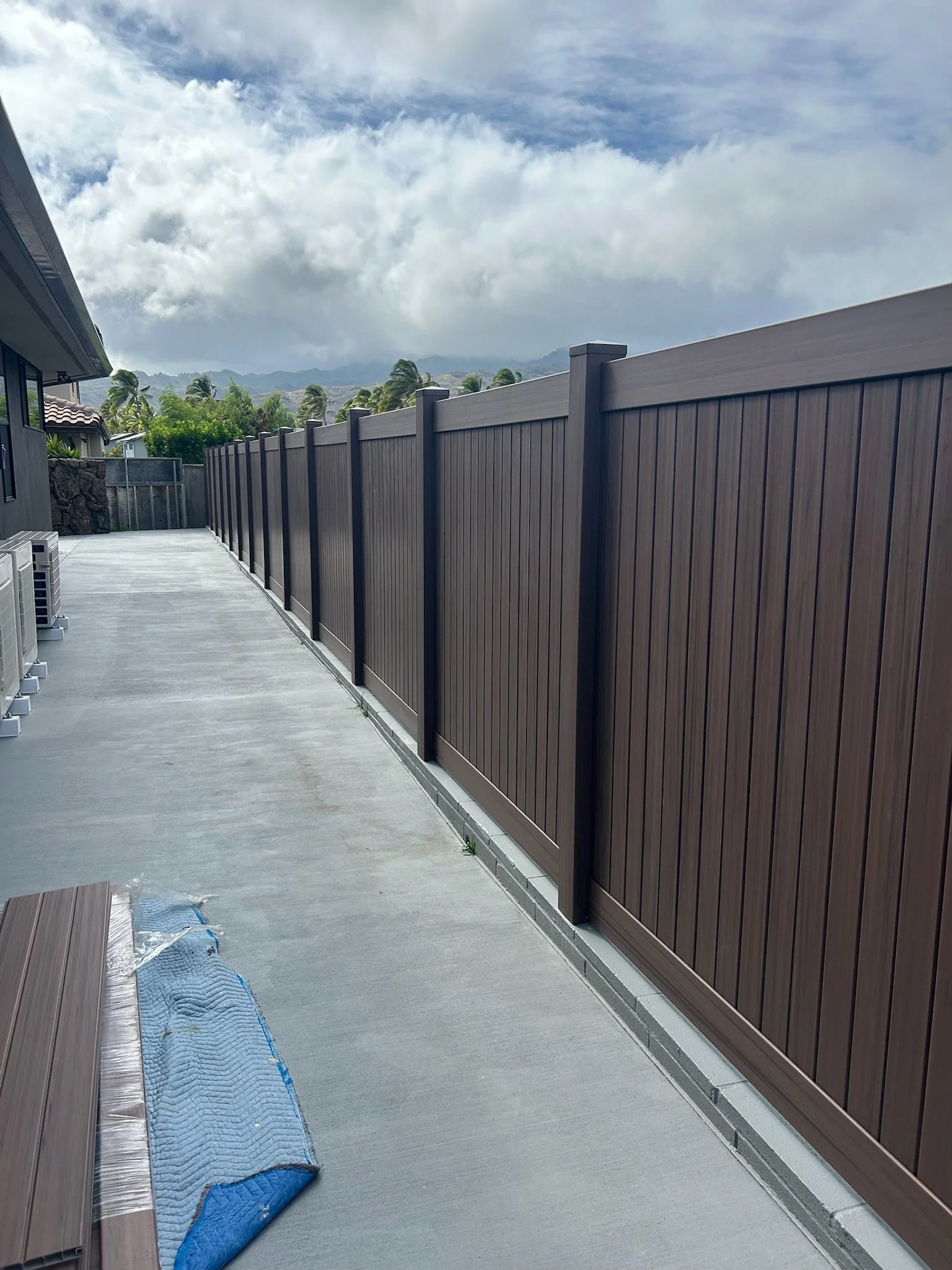 A concrete backyard patio with a brown vinyl fence on the right and a house with a tiled roof on the left. There are plants, a cloudy sky, and mountains in the distance. A blue quilted moving blanket partially covers the patio floor.