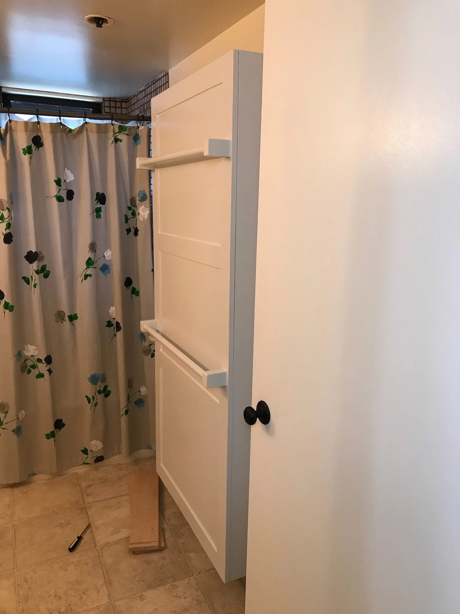 A bathroom vanity with a white cabinet, black knobs, and a black faucet, next to a beige-tiled floor, with a shower curtain featuring a floral pattern in the background.