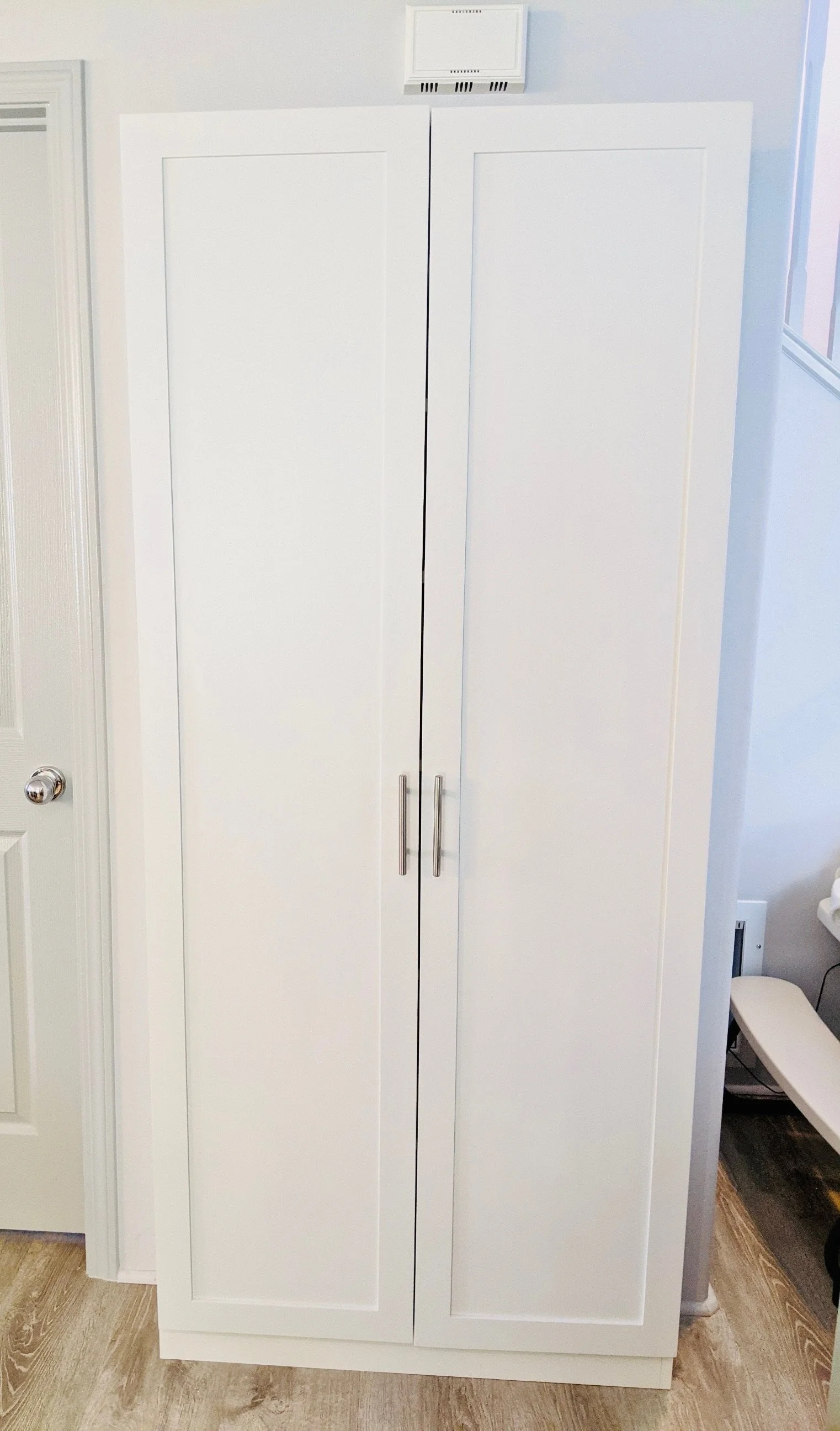 A white cabinet with two doors and silver handles, located in a room with light-colored wooden flooring, next to a door with a silver doorknob, and a vent above the cabinet.