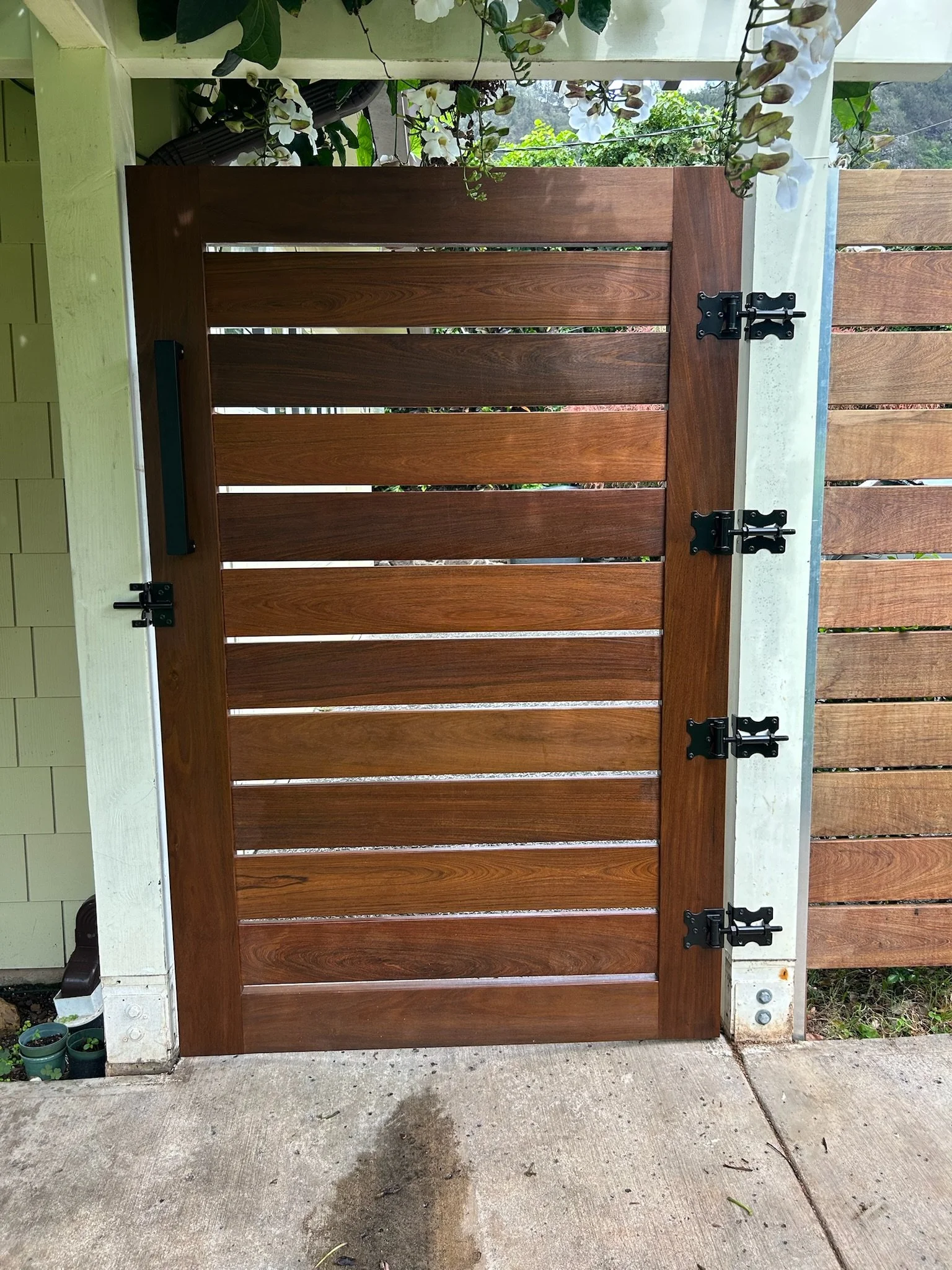 A wooden gate attached to a white fence, secured with black hinges. The gate has horizontal wooden slats and is set on a concrete ground with some small potted plants nearby. Flowering plants with white blossoms are hanging over the gate from above.