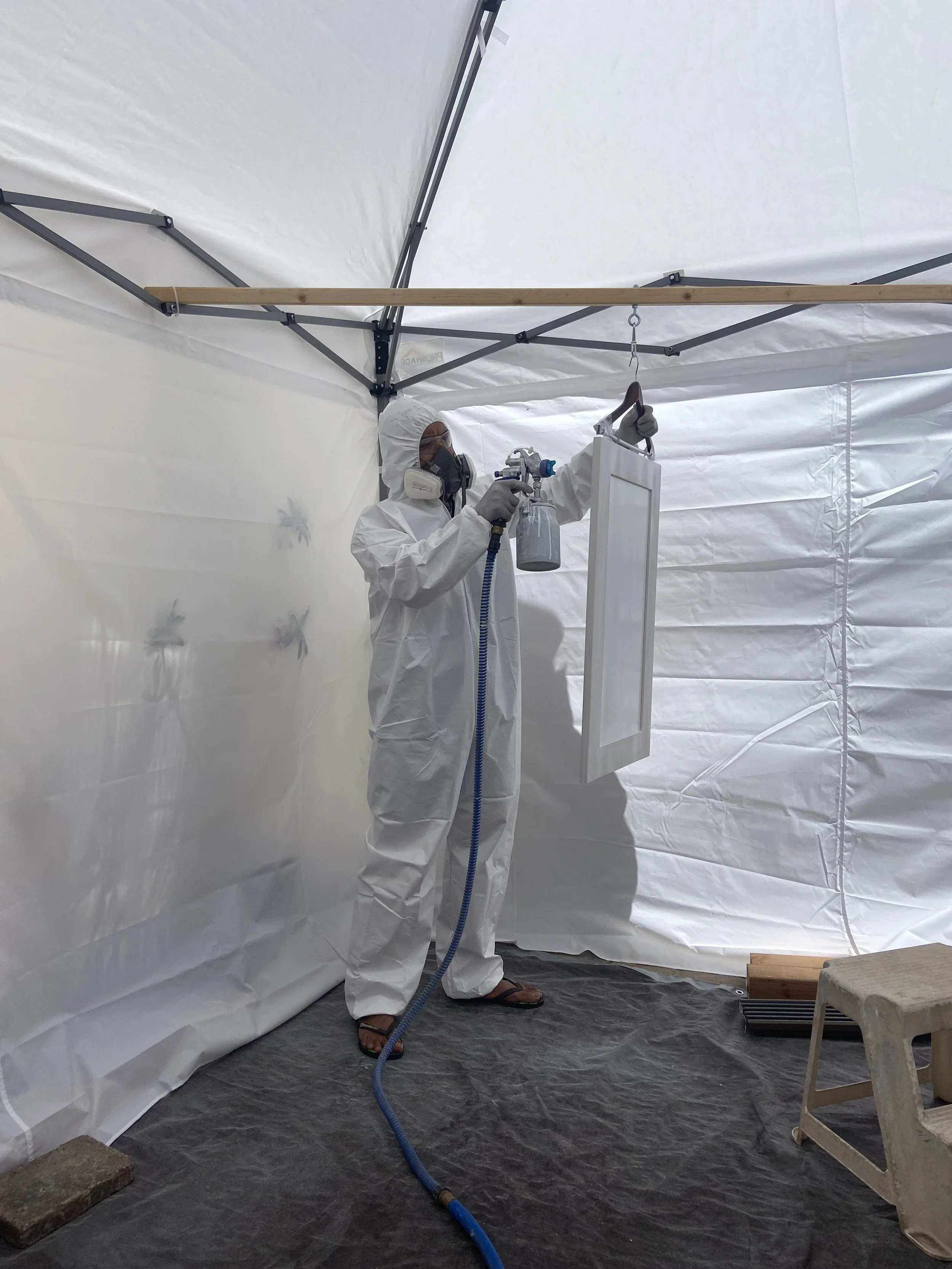 Person in protective suit, mask, and gloves spraying disinfectant inside a temporary structure with white fabric walls.