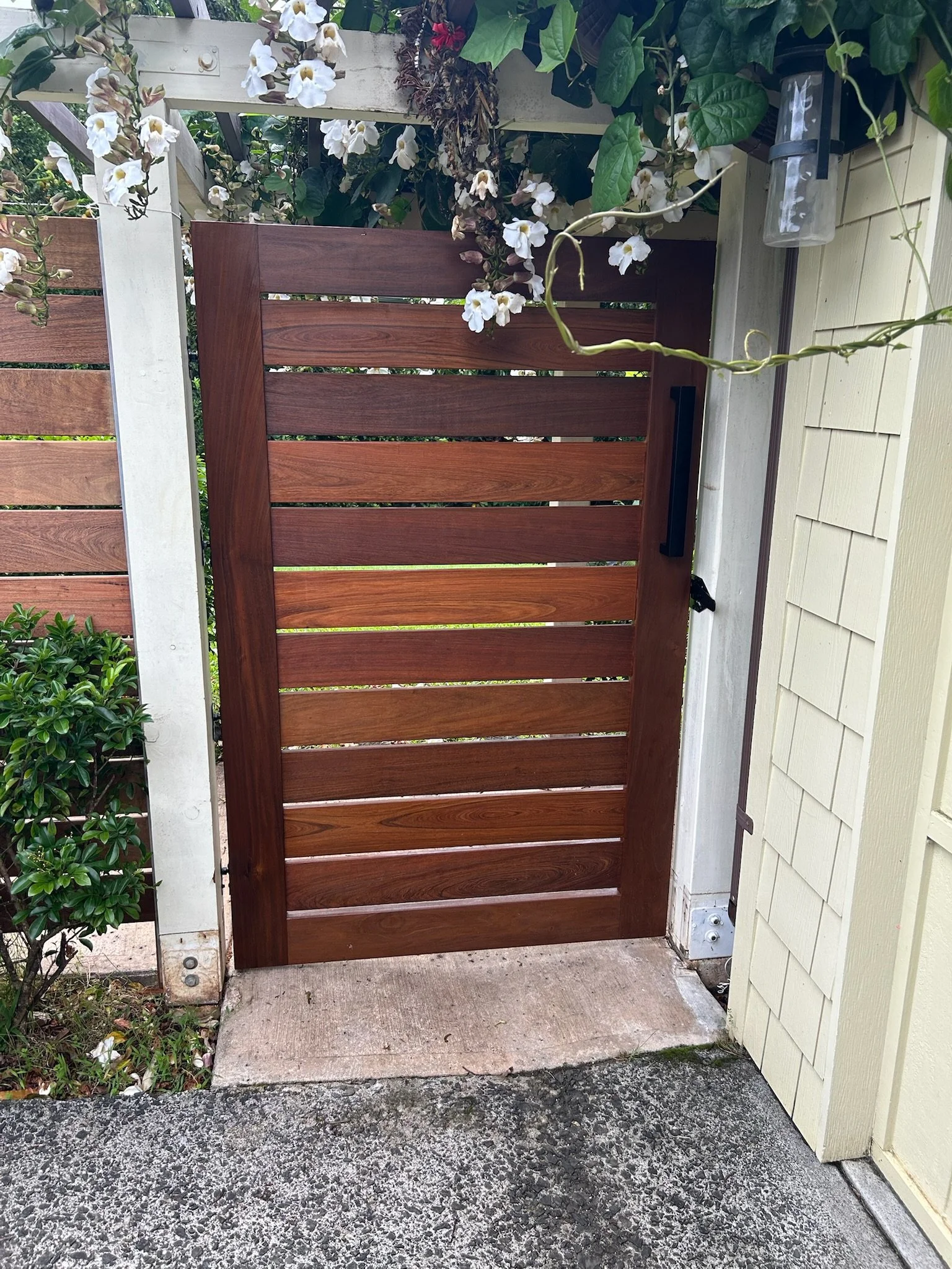 A wooden gate attached to a white fence and a beige house, with flowering plants and greenery around, on a concrete surface.