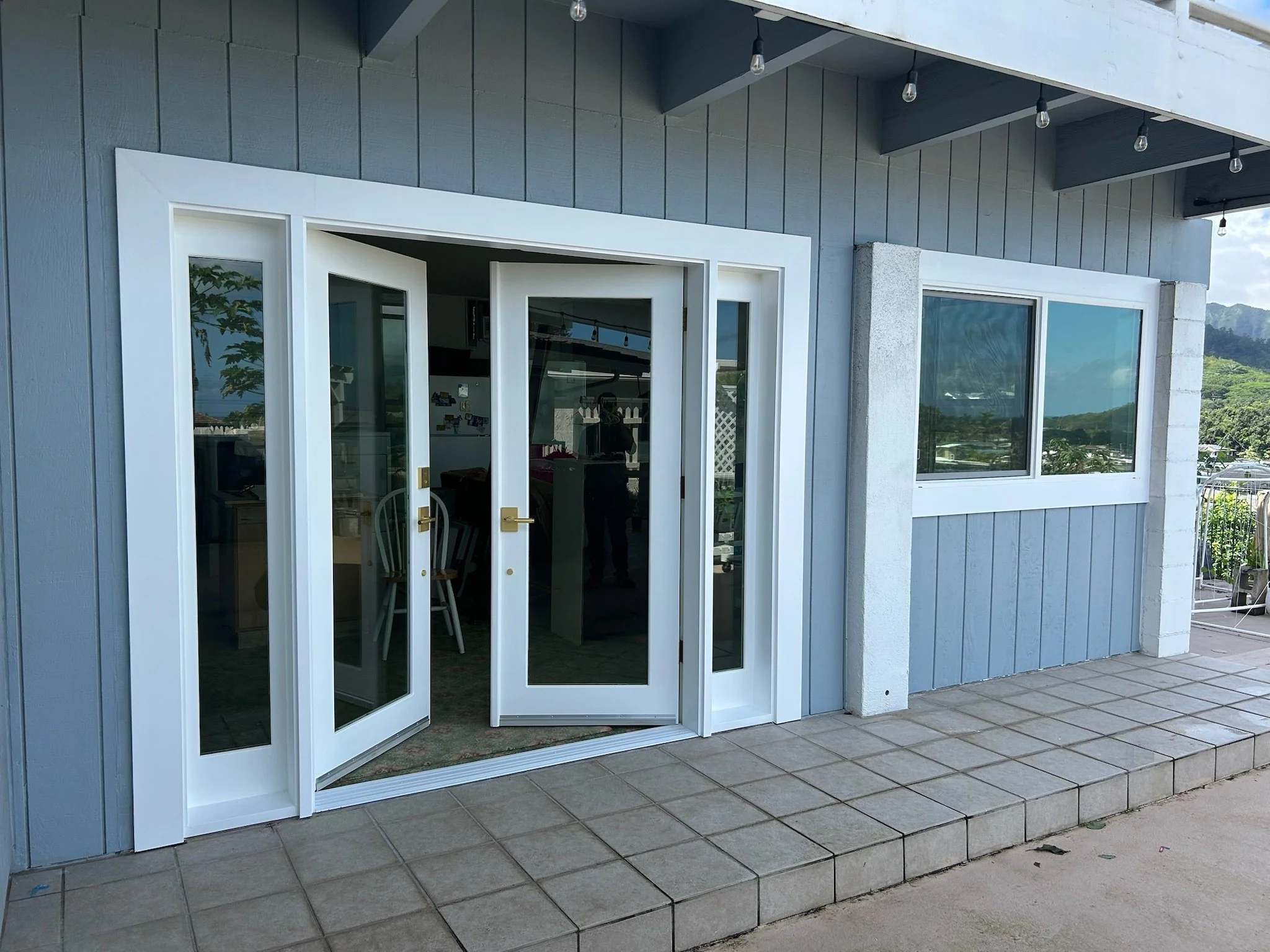 Exterior view of a house with gray siding, white window and door frames, a glass door open to reveal the interior, tiled patio, and decorative string lights overhead, with a mountain and blue sky in the background.