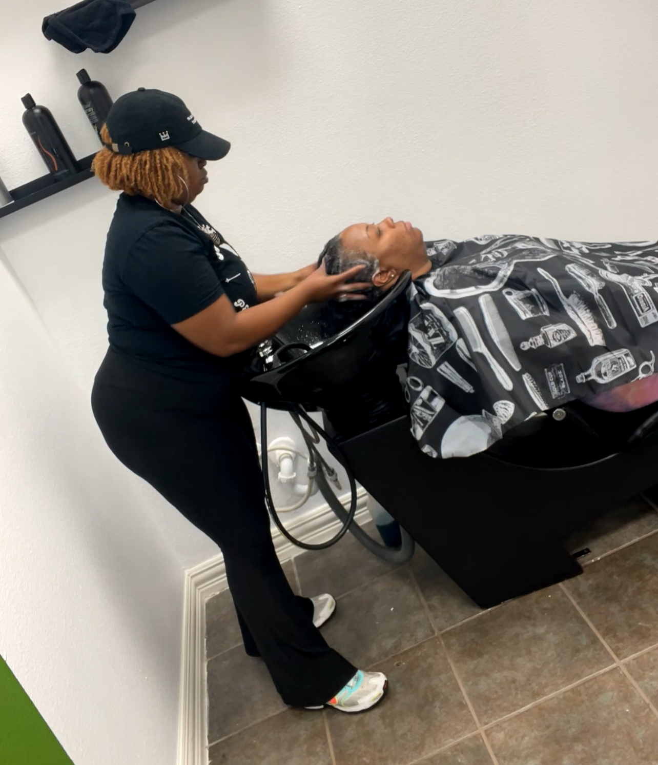 A hairstylist washing a client's hair in a salon, with the client lying back with closed eyes, wearing a patterned cape, while the stylist stands beside them.