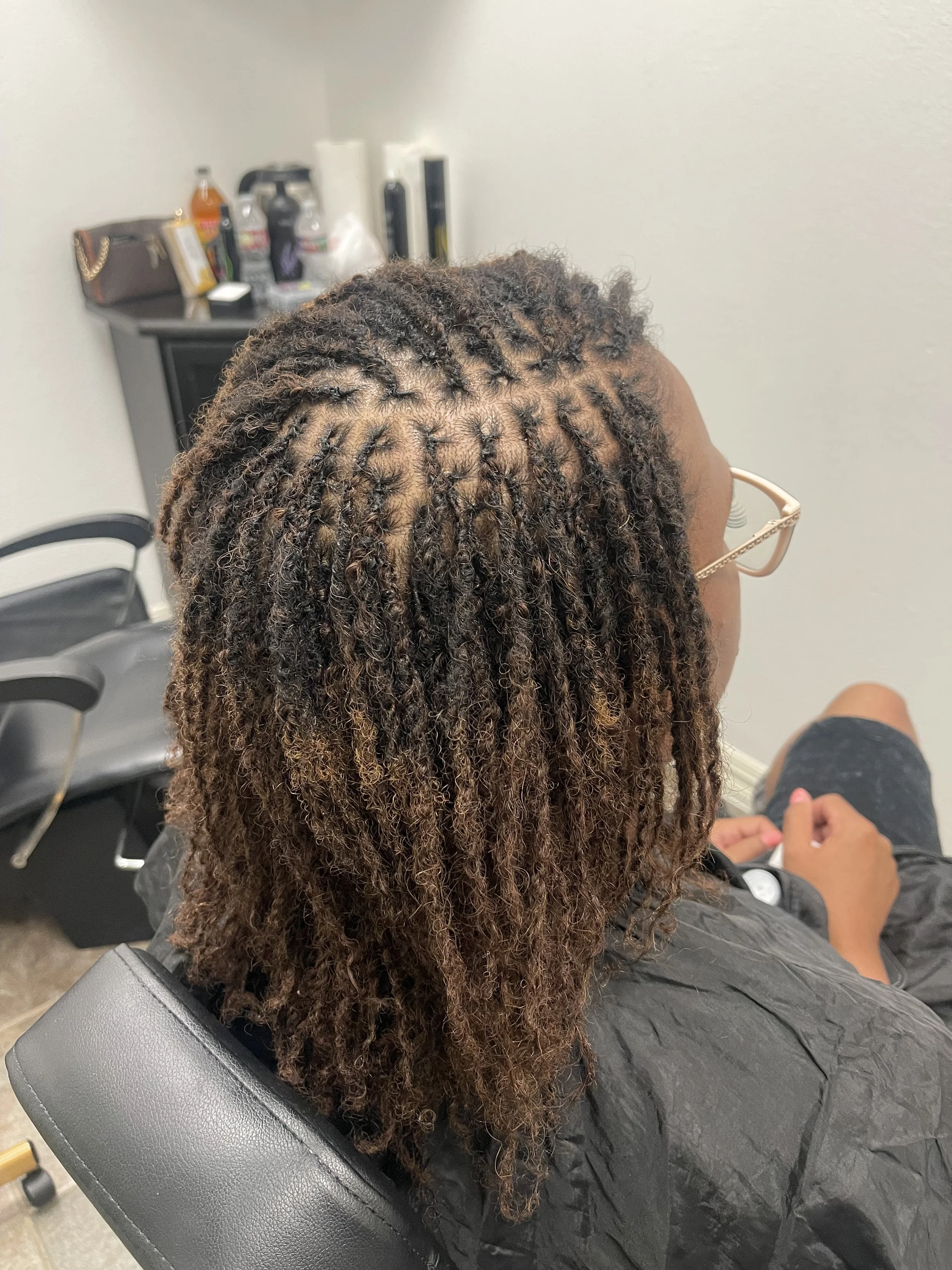 Woman with curly, dreadlocked hair sitting in a salon chair, wearing glasses and a black cape, with salon products visible on a counter behind her.