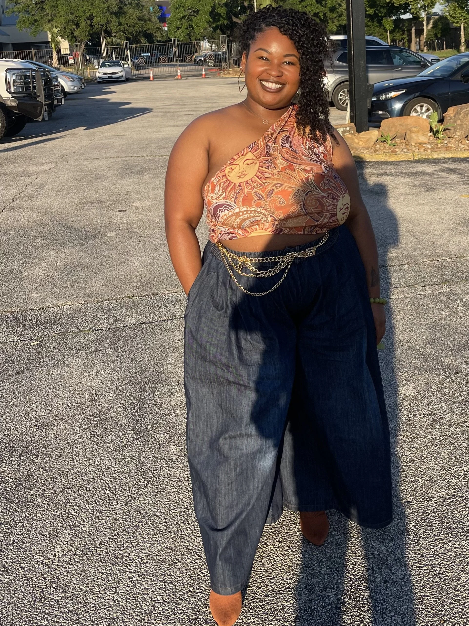 A woman standing in a parking lot during the daytime, smiling, wearing a patterned orange and brown top with sun and moon designs, dark high-waisted pants with chains, and orange shoes.