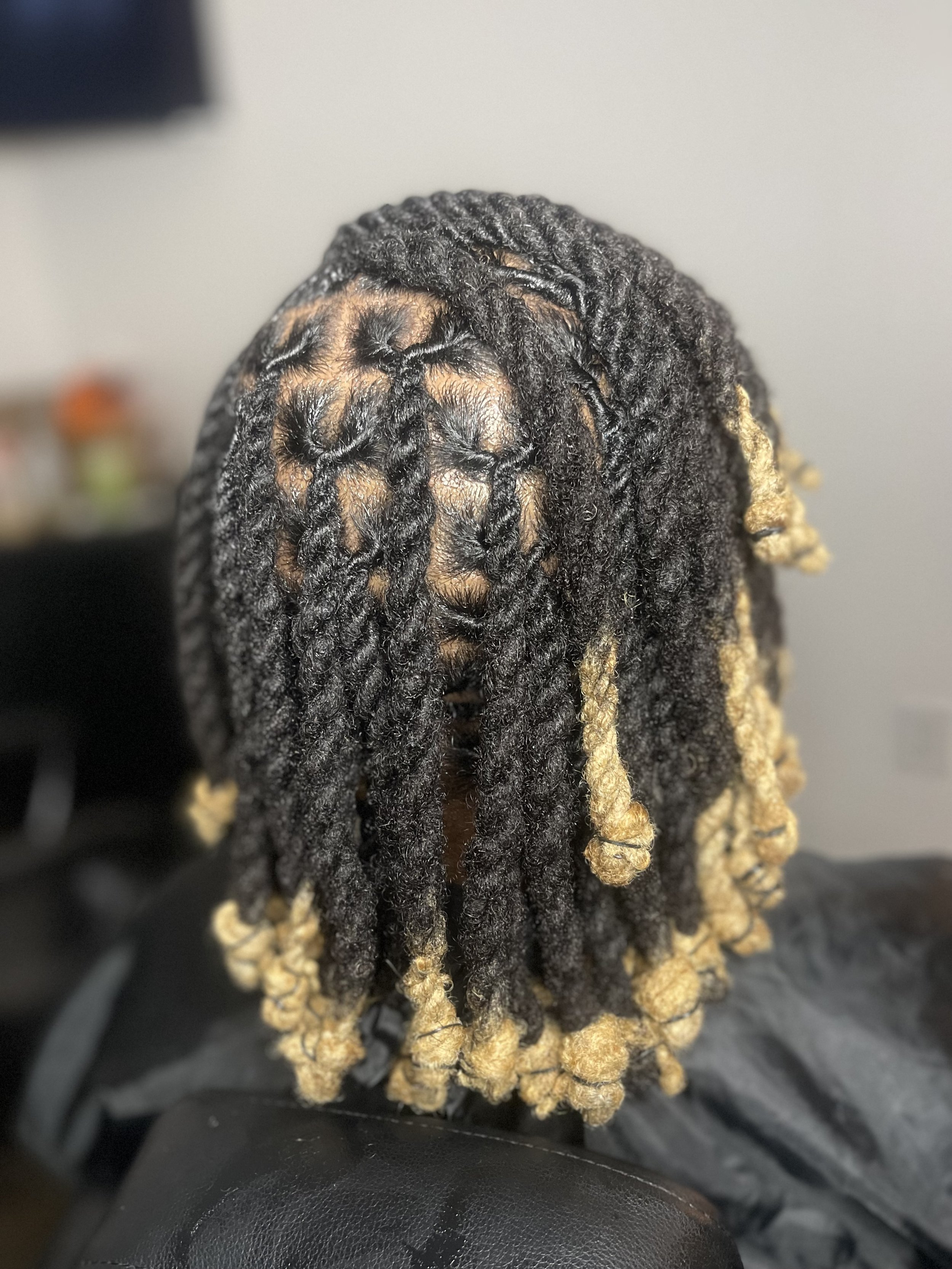 Close-up of a person's hair styled in dreadlocks with black and blond colored strands, with some dreadlocks tied at the ends, against a neutral background.