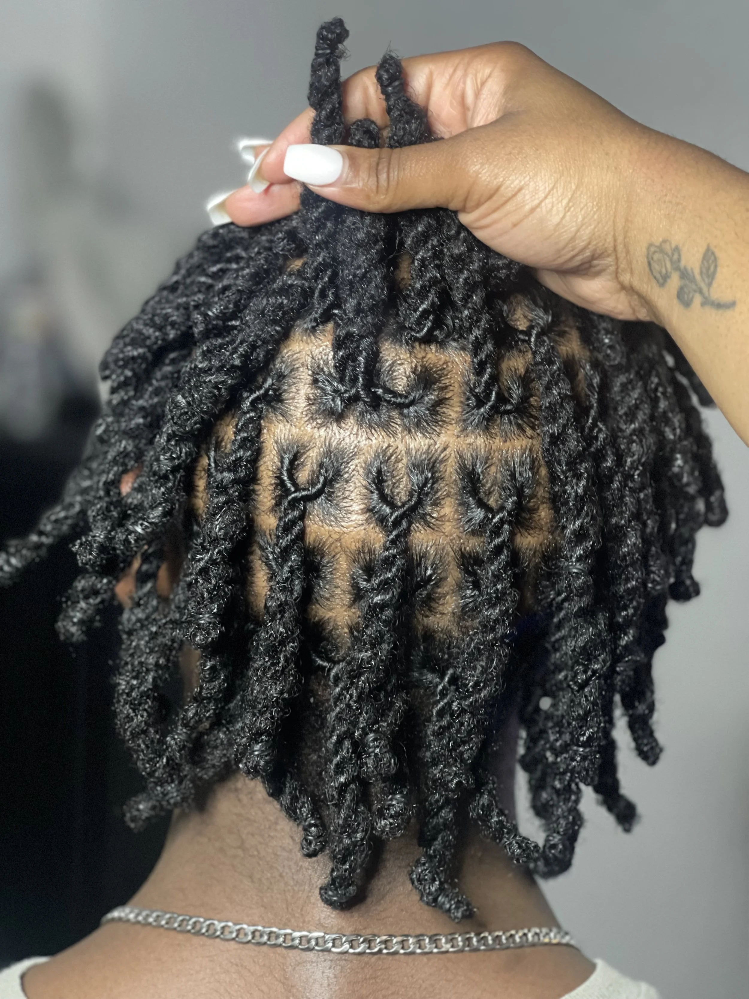 Close-up of a person's head with styled black dreadlocks held up by their hand, showing the roots and hair texture, with a silver chain necklace around their neck.