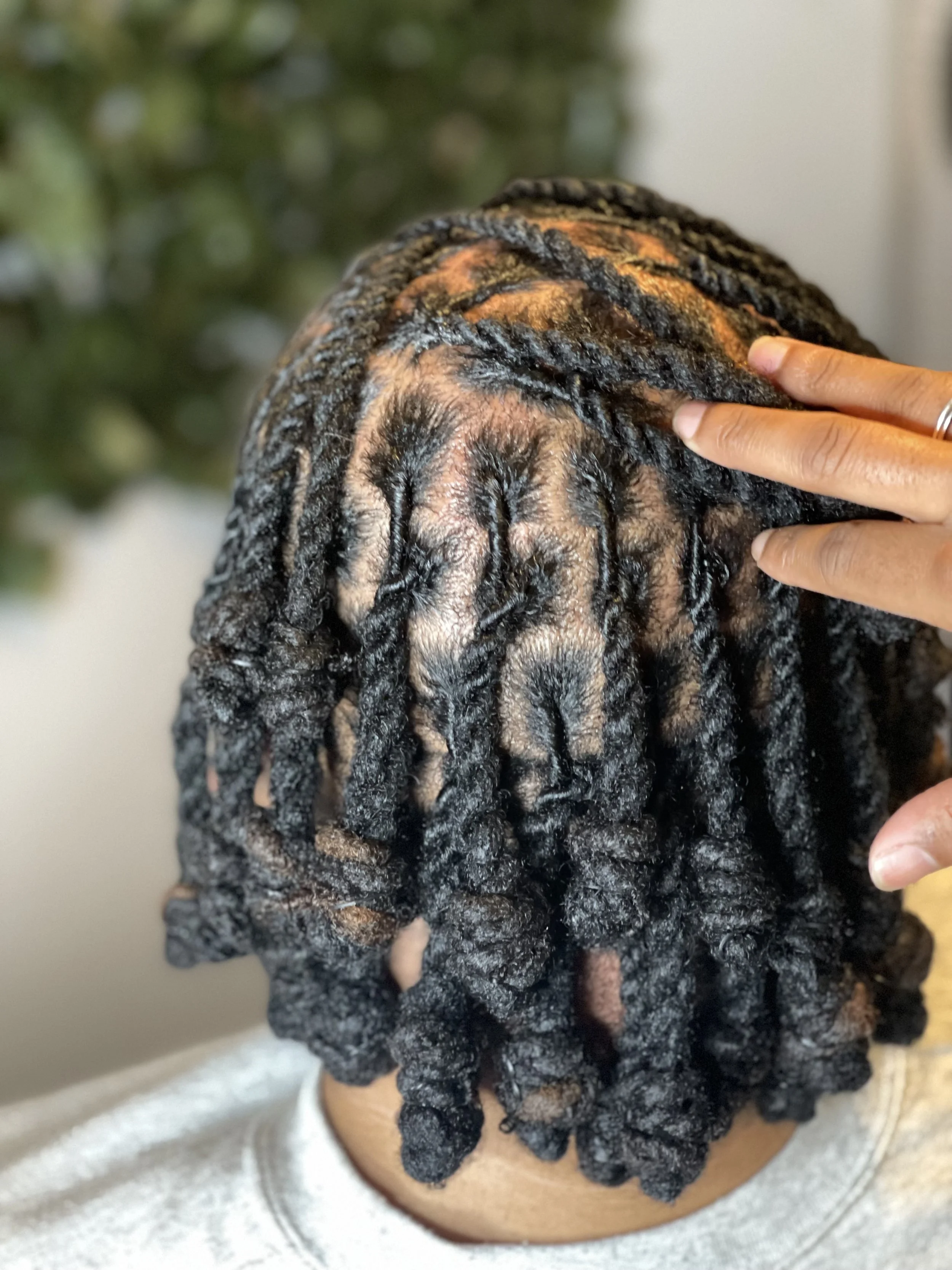 A person with braided dreadlocks touching their scalp with two fingers, with a blurred background of green foliage.