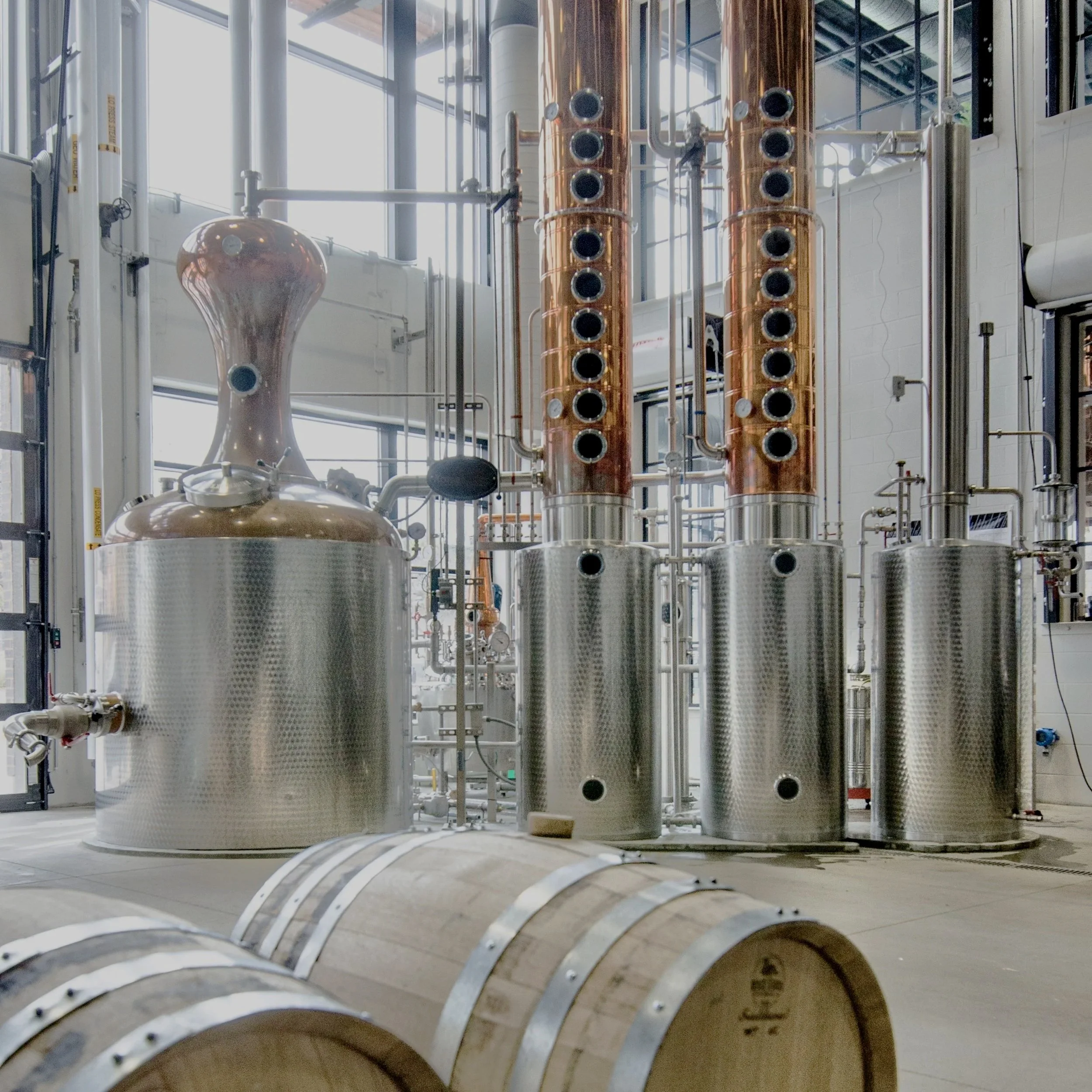 Industrial brewery equipment with copper and stainless steel tanks, pipes, gauges, and cooling coils inside a modern facility.