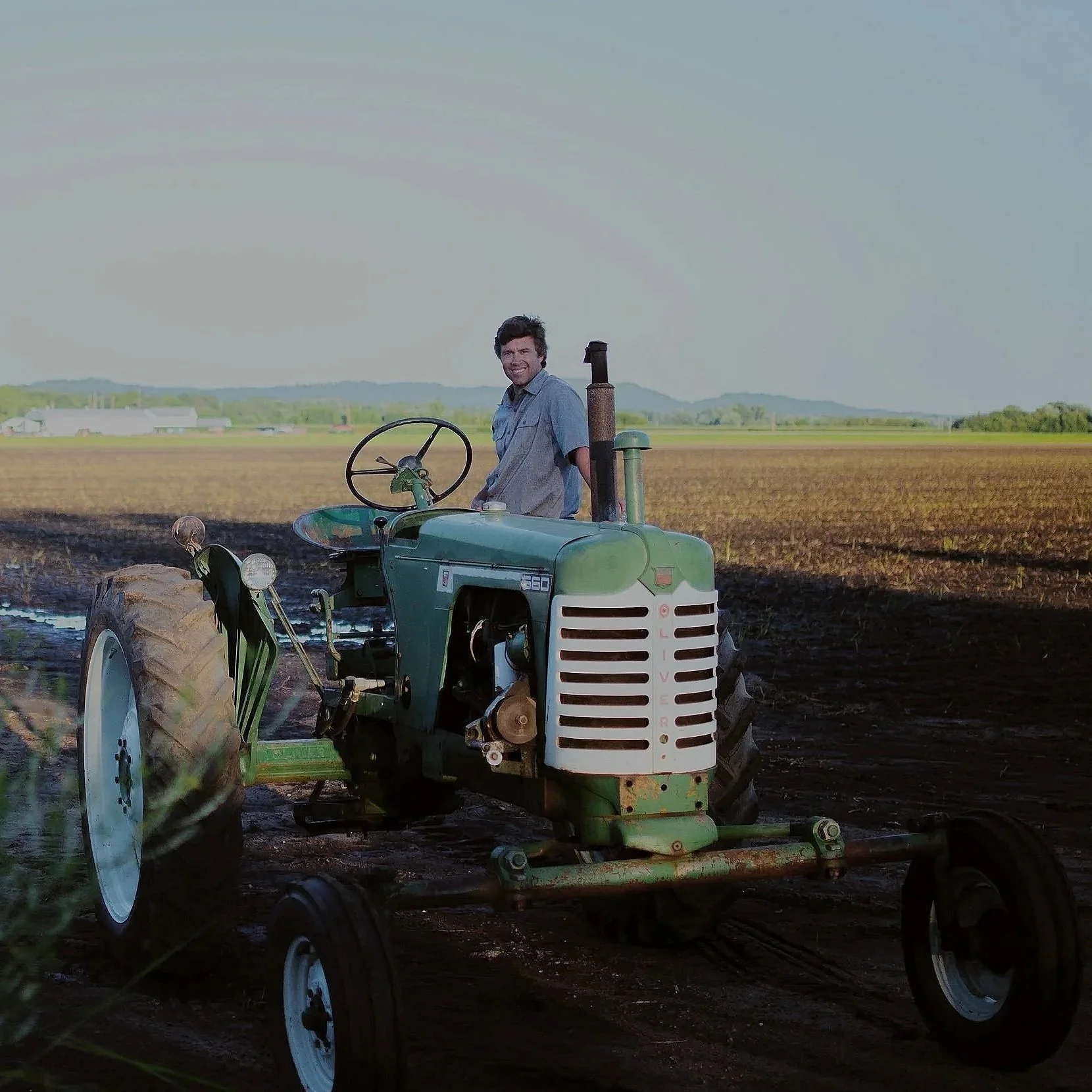 A man standing on a vintage green tractor in a rural farm field under a cloudy sky, smiling with trees and buildings in the background.