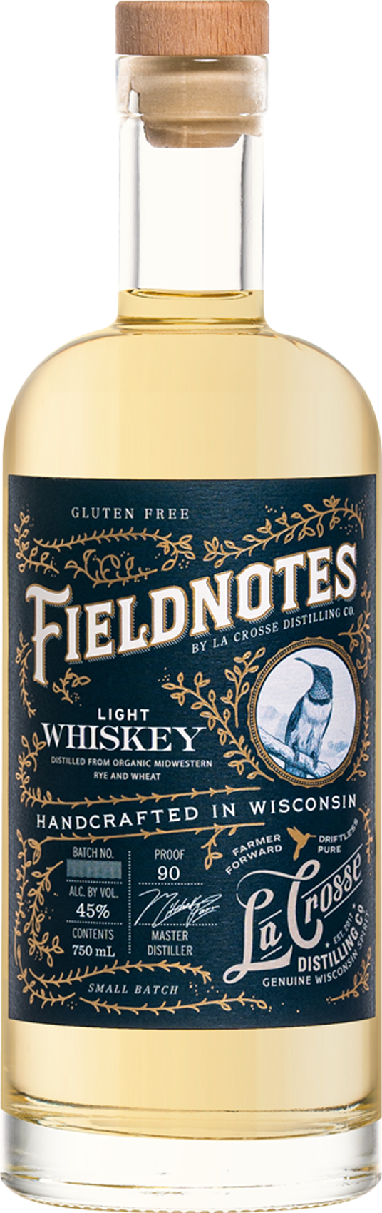 A clear glass bottle of Fieldotes light whiskey with a wooden stopper and a dark blue label featuring gold and white decorative text and designs, including a bird illustration, indicating it is gluten-free, handcrafted in Wisconsin, and distilled from organic Midwestern rye and wheat.