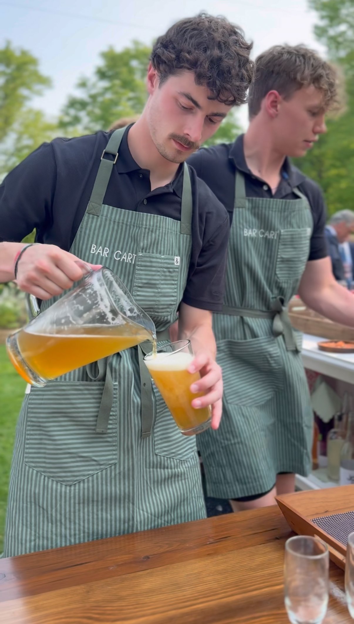 Two young men wearing striped aprons pouring beer into glasses at an outdoor gathering.