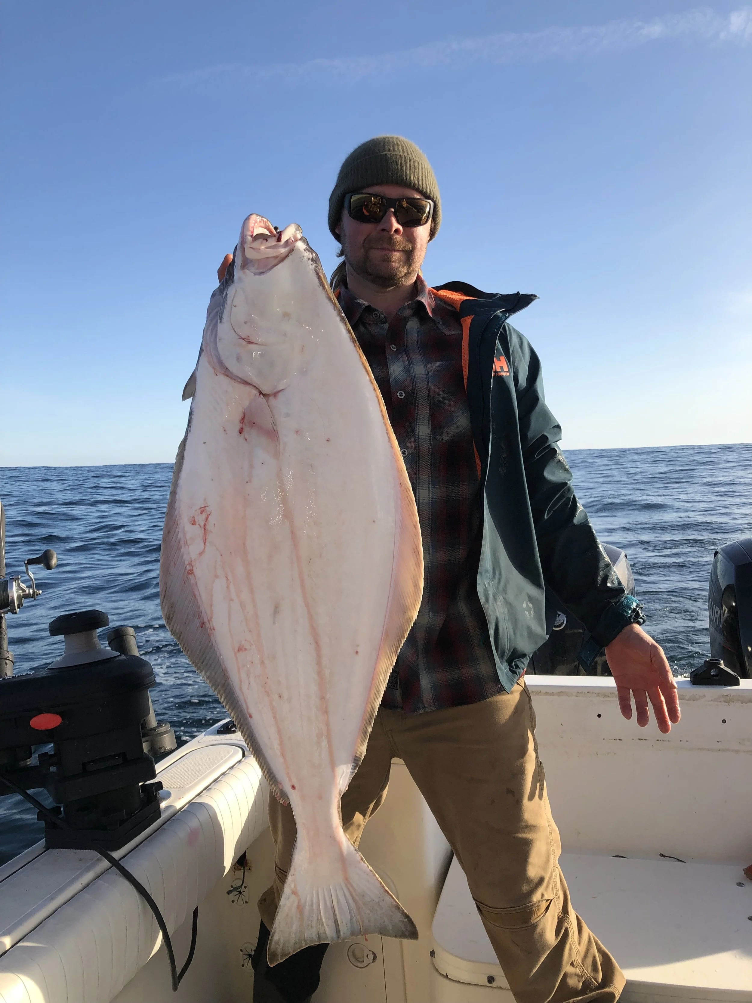 A man wearing sunglasses, a knit beanie, and outdoor gear holding a large fish on a boat at sea during daylight.
