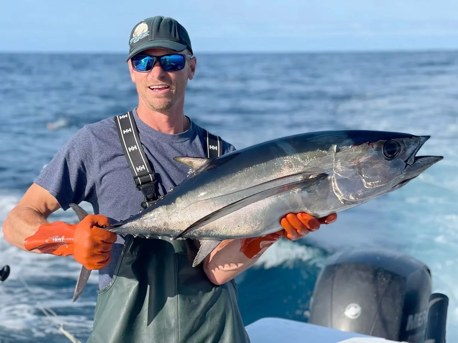 A man wearing sunglasses, a hat, and orange gloves holds a large fish on a boat in the ocean with a blue sky in the background.