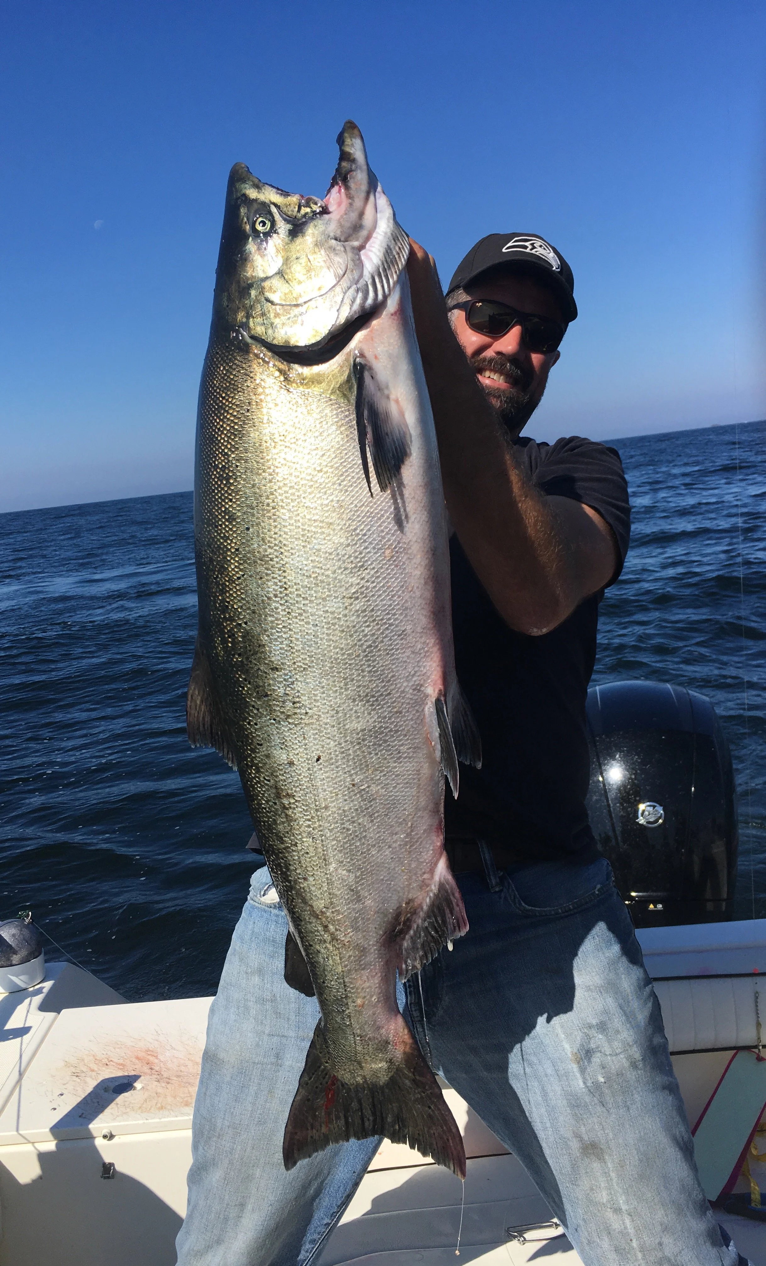 A man holding a large fish on a boat in the open ocean, wearing sunglasses and a black cap.