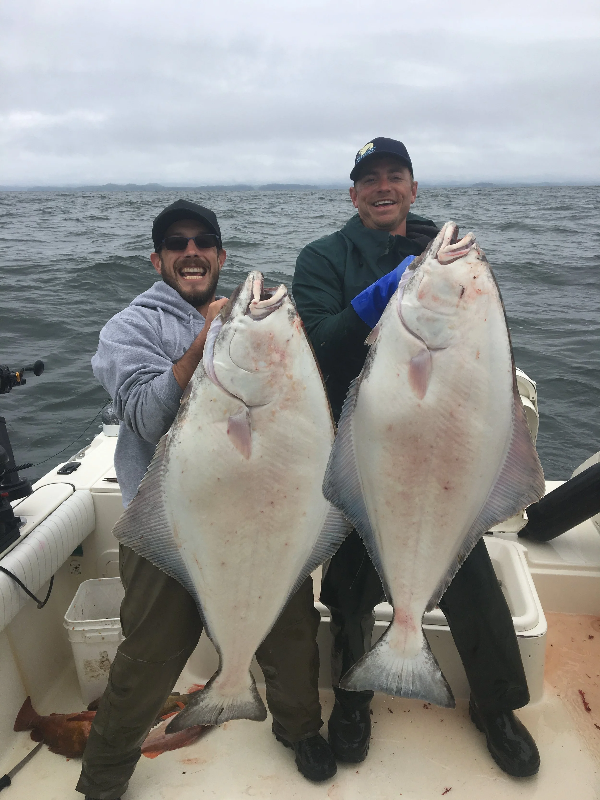Two men on a boat holding large halibut fish, smiling, overcast sky in the background.