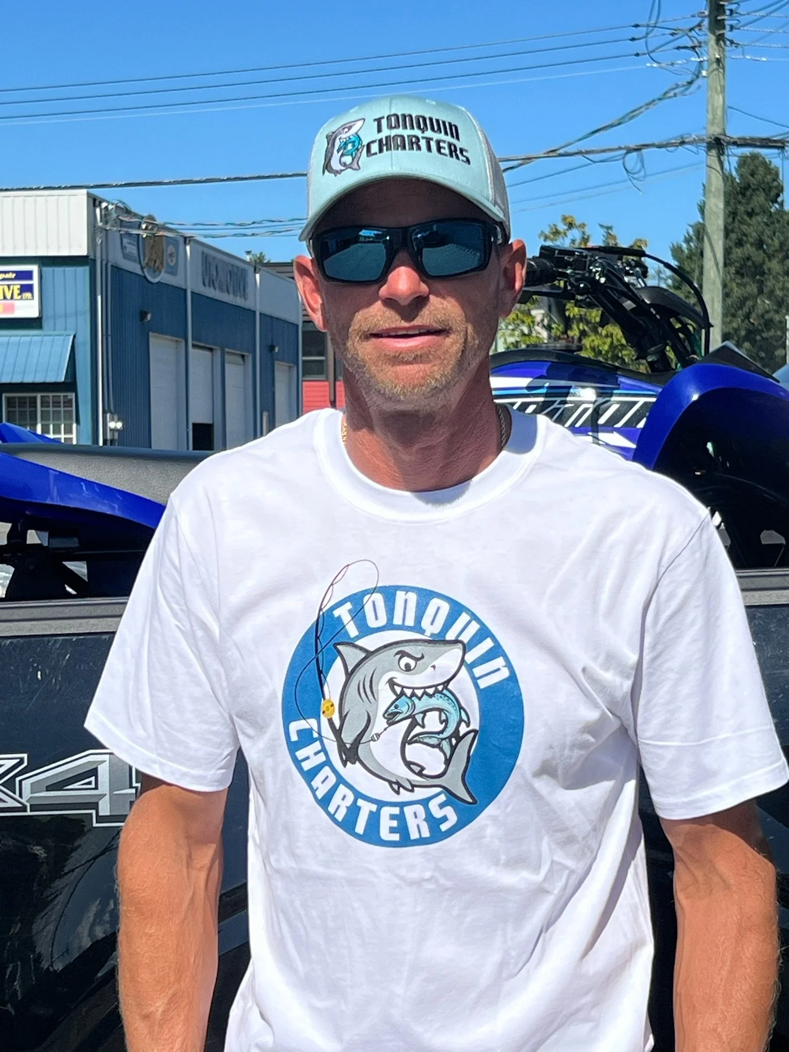 A man wearing a white T-shirt and a baseball cap with the 'Tongue Cutters' logo, standing outdoors in front of a blue building, power lines, and a blue vehicle.