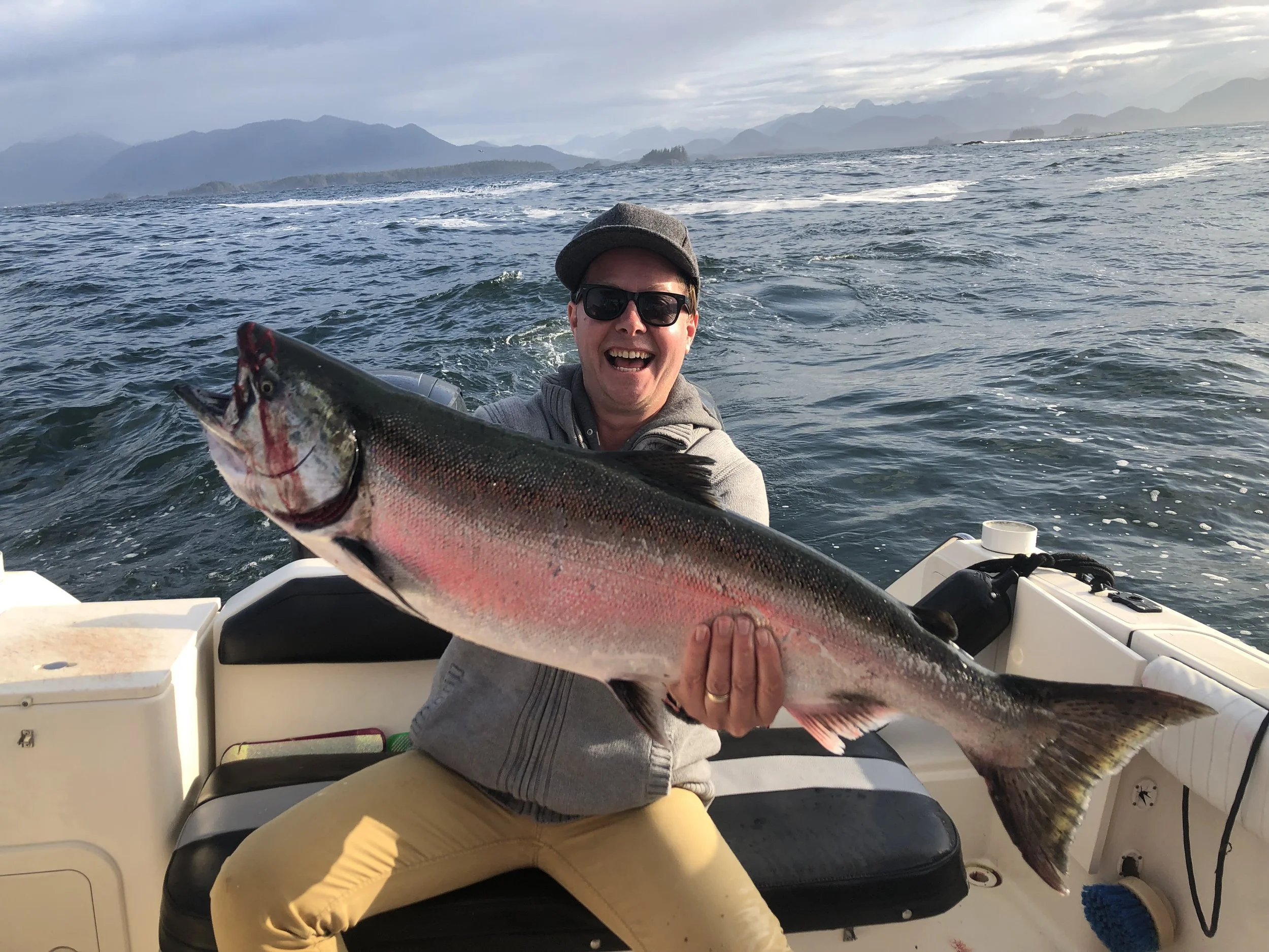 Man on boat holding a large fish with a mountainous background and cloudy sky.