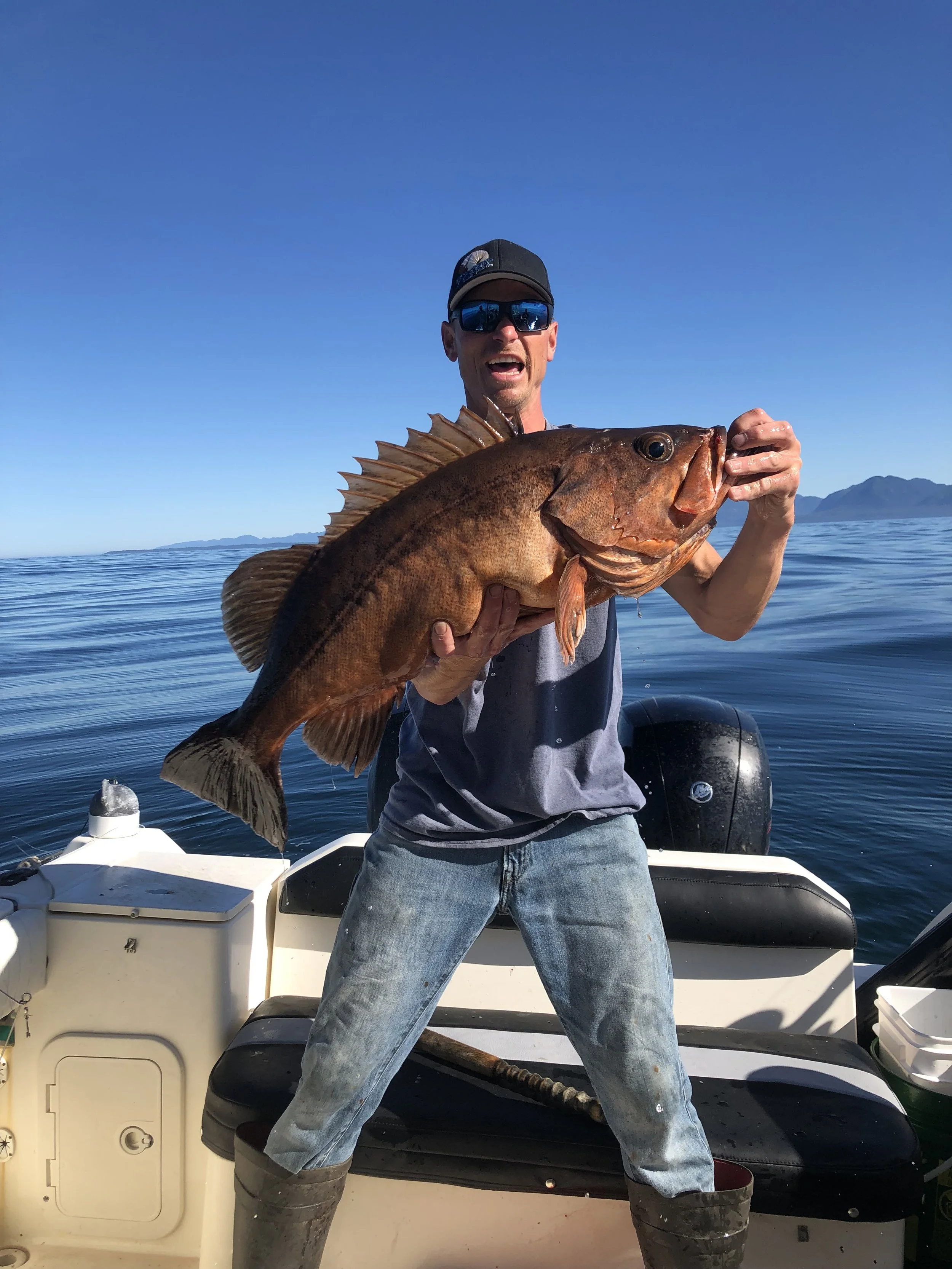 Man holding a large brown fish on a boat, with a background of calm water, mountains, and a clear blue sky.