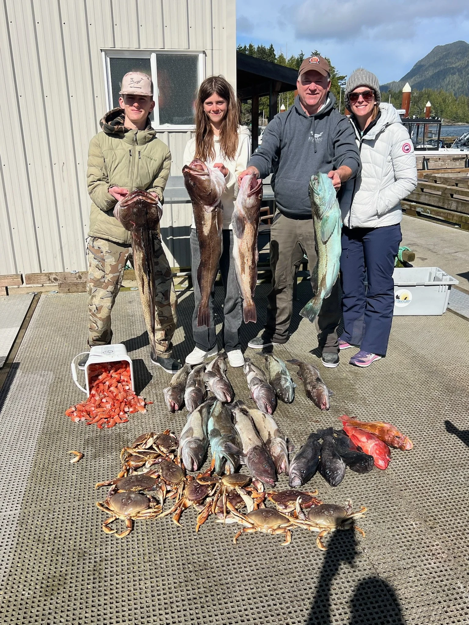 Group of four people on a dock holding large fish. Fish and seafood laid out on the dock in front, including crabs, lobster, and various fish. A container of chopped seafood is nearby. Background features mountains, water, boats, and cloudy sky.