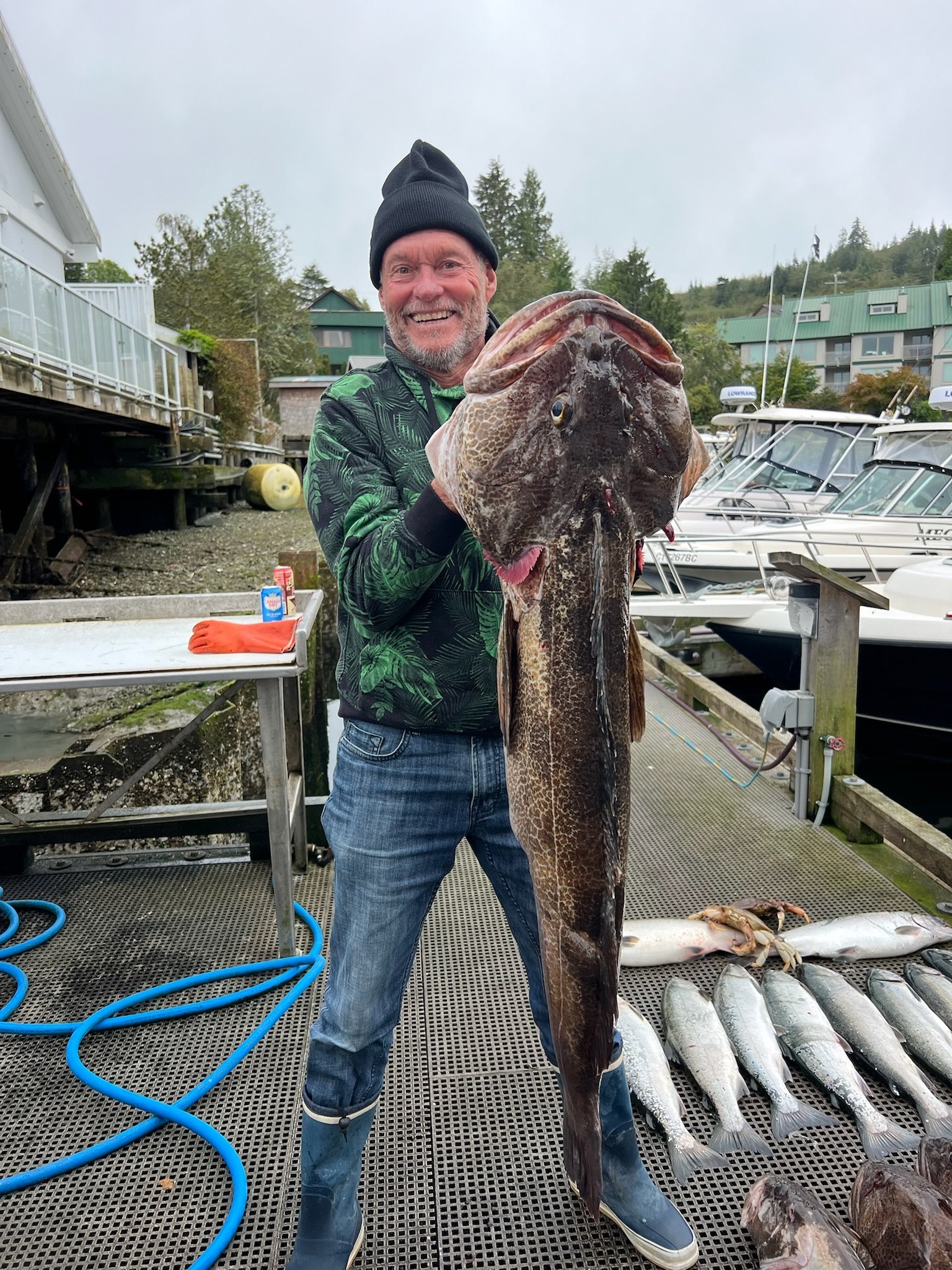 A man in rain boots and a hoodie holding a large fish on a dock with boats and smaller fish lying on the ground.