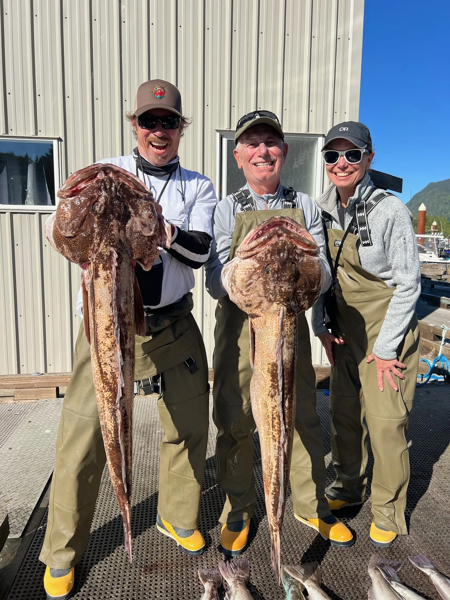 Three people smiling and holding large fish in front of a metal building on a boat deck, with mountains and blue sky in the background.