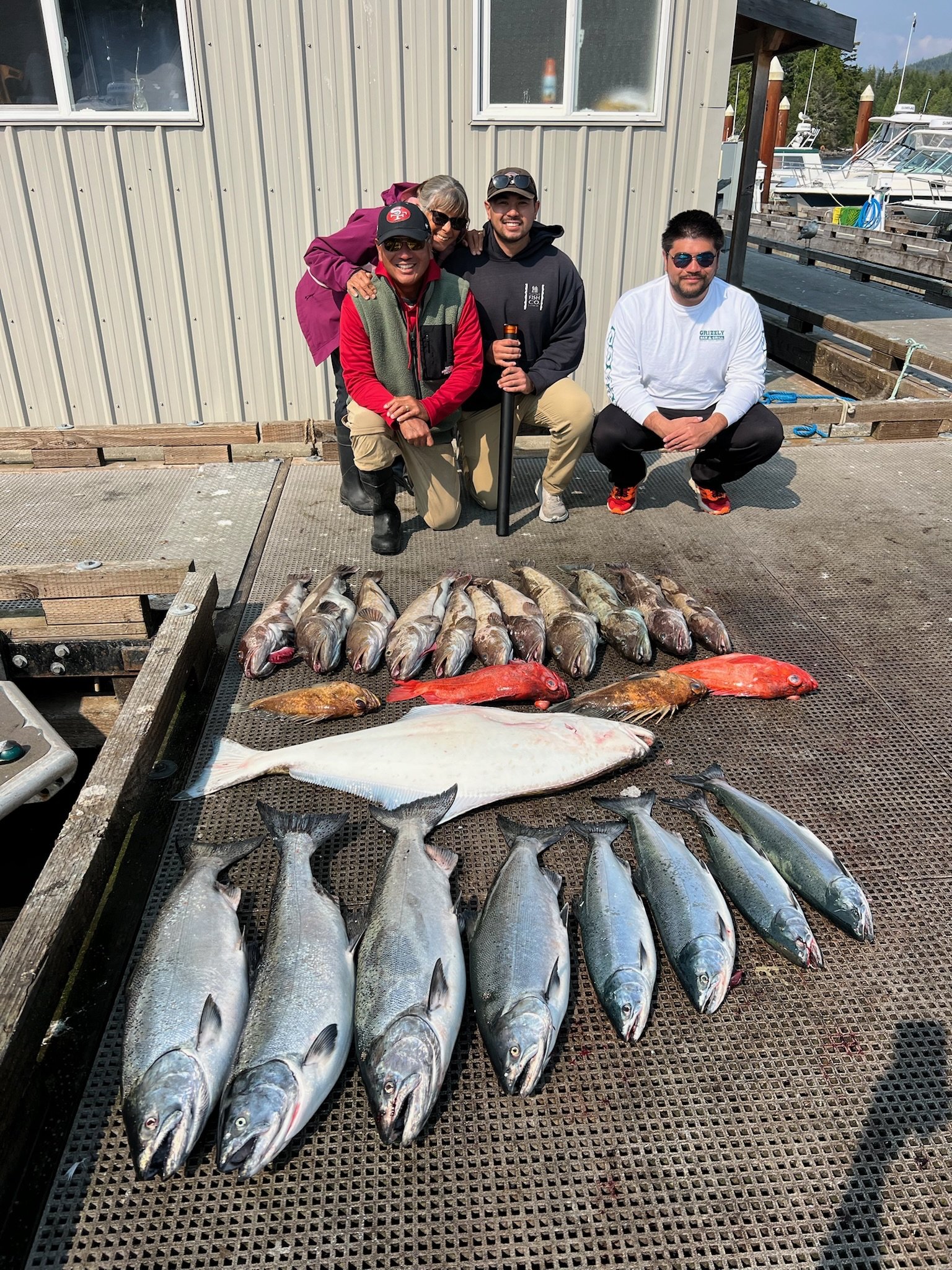 Group of five men kneeling and squatting behind a large catch of fish displayed on a platform outside near a marina, with boats docked in the background.