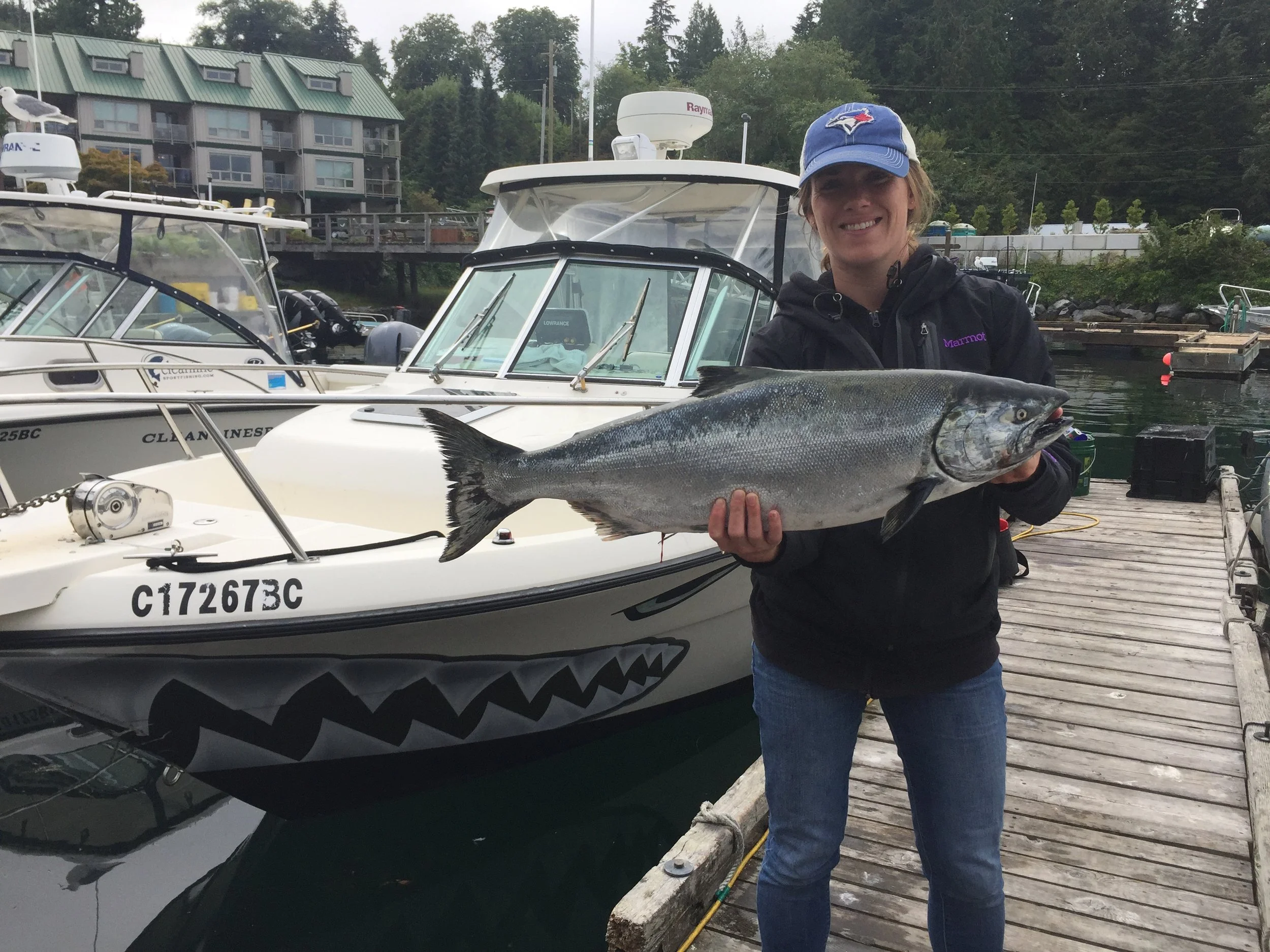 Woman wearing a blue cap with a Toronto Blue Jays logo and a black jacket, smiling and holding a large fish near boats docked at a marina, with trees and buildings in the background.
