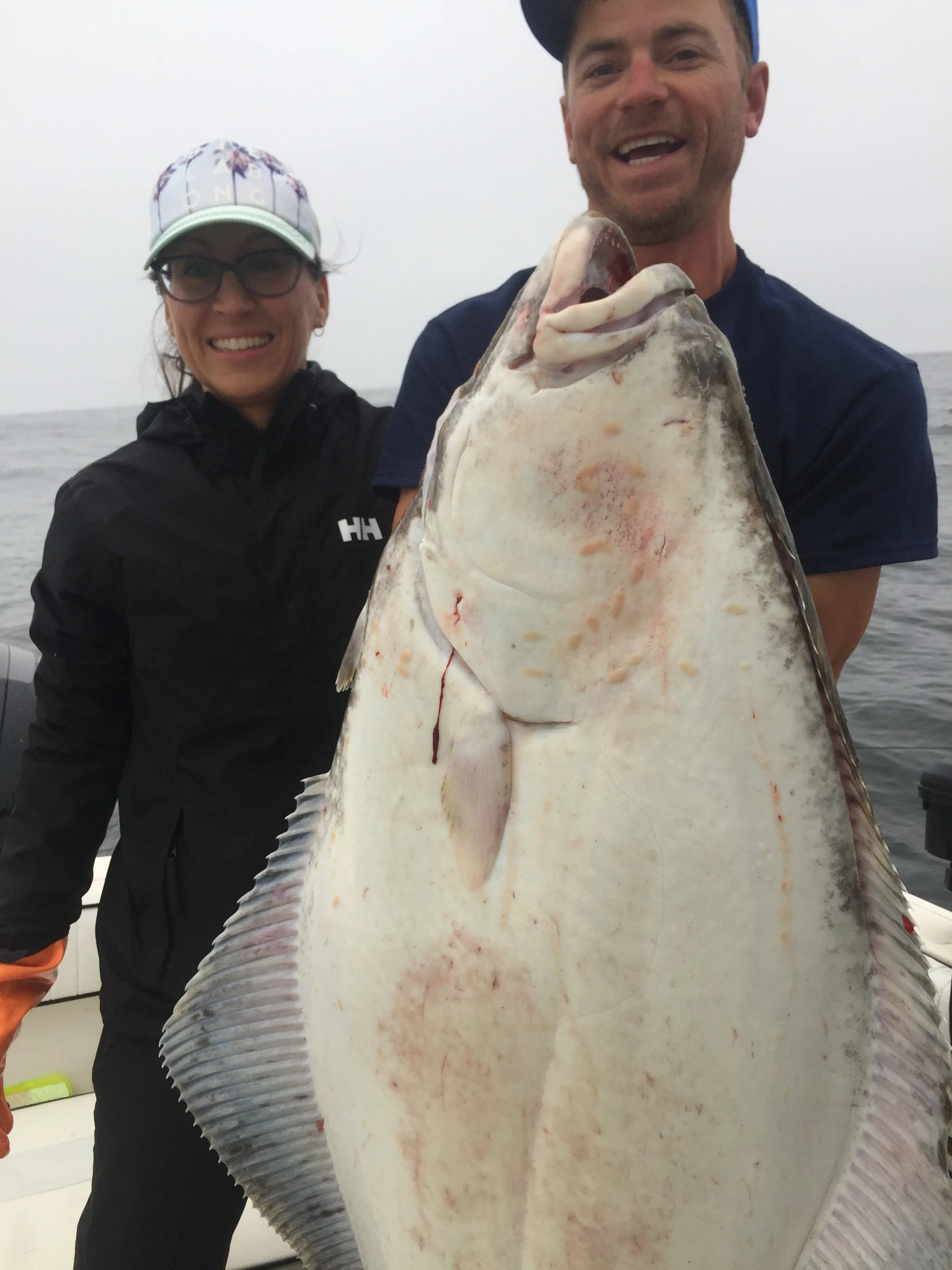 Two people smiling on a boat holding a large halibut fish.