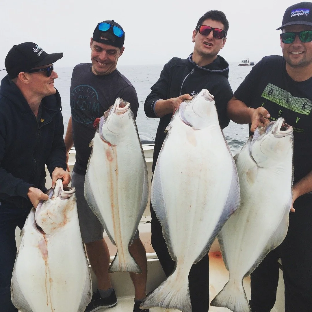 Four men on a boat holding large halibut fish they caught, smiling and wearing sunglasses with a foggy sky and water in the background.